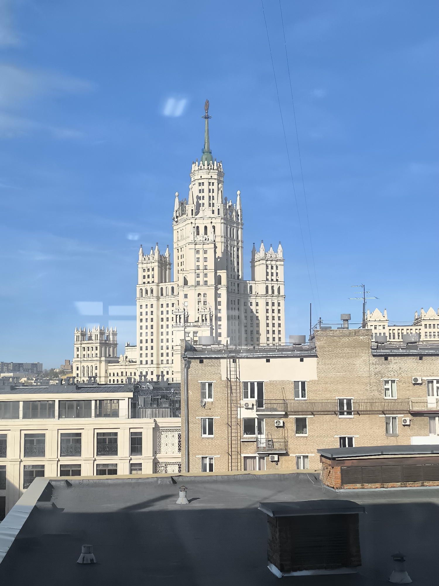 The image captures a vibrant daytime cityscape in Moscow, Russia, dominated by a magnificent Stalinist-era high-rise building. This ornate, light-colored edifice, reminiscent of the "Seven Sisters," features multiple tiers, classical architectural elements, and a tall, slender spire capped with a golden-hued ornament. Sunlight illuminates its intricate facade and numerous windows, giving it a bright appearance against a clear, deep blue sky.
In the mid-ground and foreground, a collection of other urban structures is visible. To the left of the main building, another, slightly smaller but similarly styled building stands. Closer in the foreground are several lower-rise buildings with flat roofs, some featuring brick facades and others with lighter-colored walls. These roofs are adorned with various industrial elements such as vents, chimneys, air conditioning units, and ladder access points. A section of a brick building in the lower right shows multiple windows and external air conditioning units, some with small balconies.
The sky is largely cloudless, save for a few very faint, wispy patches and some prominent rectangular light reflections, suggesting the photograph was taken through a window. Thin, dark electrical wires or cables stretch diagonally across the upper portion of the sky. The bright sunlight indicates it is either morning or early afternoon on a clear, pleasant day. There is no discernible activity or specific event taking place, and no visible text in the image.