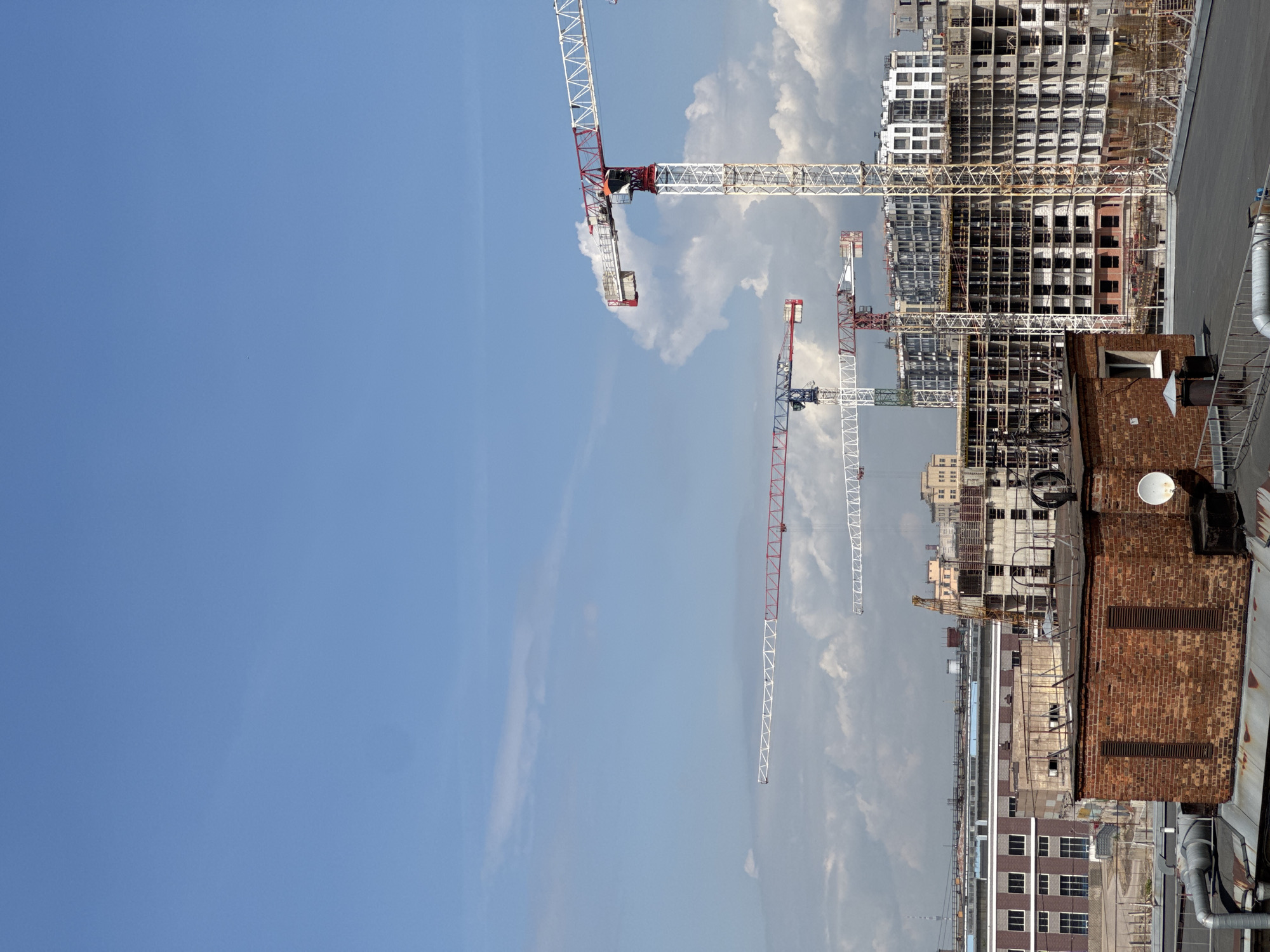 This media file captures an expansive view of an urban construction site under a partly cloudy blue sky. The scene is dominated by several large tower cranes and multiple buildings in various stages of construction, suggesting ongoing development.
In the upper and mid-ground, at least three prominent tower cranes are visible. The closest one features a white and red frame with its long boom extending across the frame. Further back and to the right, other cranes, also white with red or blue accents, are positioned among the rising structures.
The right side of the image features numerous multi-story buildings under construction. These appear to be modern high-rises, with their concrete frameworks, exposed rebar, and empty window frames clearly visible. Some buildings show early facade work, while others are just structural skeletons. Towards the foreground on the far right, an older, brick building with a flat roof, vents, and a white dish-like object provides a contrast to the new construction.
The sky occupies a significant portion of the left and upper parts of the image, showcasing a clear blue with scattered, fluffy white clouds. The lighting suggests it is daytime, possibly midday or early afternoon, with no strong shadows indicating a particular angle of the sun. No people are visible within the frame. There is no visible text or specific architectural cues to definitively identify the exact location. The overall atmosphere conveys a sense of growth and urban expansion.
