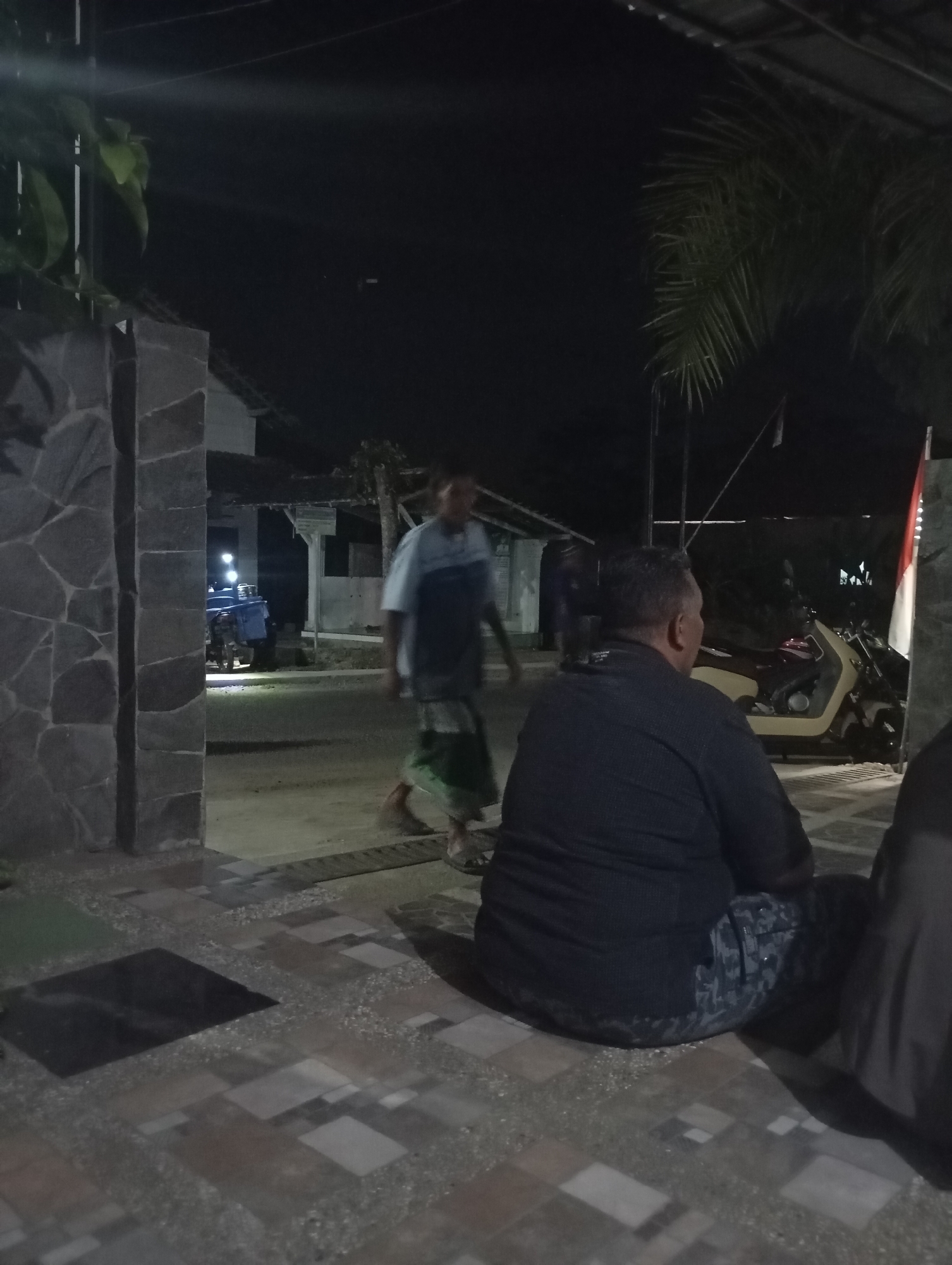 A medium shot captures an outdoor scene at night, possibly in Banjarsari, Indonesia, focused on a man sitting and looking away from the camera. He is wearing a dark, collared shirt and patterned pants. To his left, a motorcycle is parked. Further to the left, a man in a light blue shirt and green patterned garment walks away from the camera, his face obscured. In the background, a building with a tiled roof and a white sign is visible. The ground is paved with patterned tiles, and a stone wall runs along the left side of the frame. A palm tree is visible on the right side of the image, and a flag, possibly the Indonesian flag, is mounted on a pole. The lighting suggests artificial light sources.