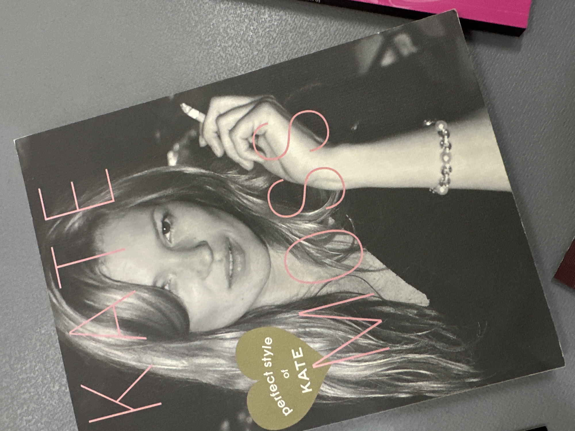 The image shows a book cover featuring a black-and-white photograph of Kate Moss. She is holding a cigarette in her right hand, and a silver bracelet is visible on her wrist. Pink lettering spells out "KATE MOSS" on the cover. A golden heart-shaped graphic reads "perfect style of KATE." The image appears to be taken indoors. There's no information indicating the location of Tunbridge Wells, United Kingdom.