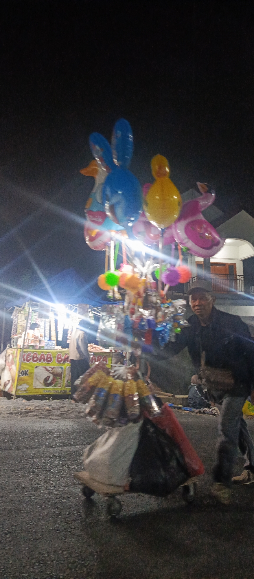 A street vendor stands near a cart filled with various items, including balloons shaped like animals and small toys. The vendor, a man wearing a dark jacket and a baseball cap, is looking towards the camera with a slight smile. The cart is positioned on a paved street at night. In the background, there is a food stall with a banner that reads "KEBAB BAK SO", indicating a kebab food business. Further back, a building with a white facade and a red-tiled roof is visible. The overall scene appears to be a night market or street-side stall area in Sumbersekar, Indonesia. The lighting suggests artificial illumination from the stalls and potentially streetlights, casting some lens flares.