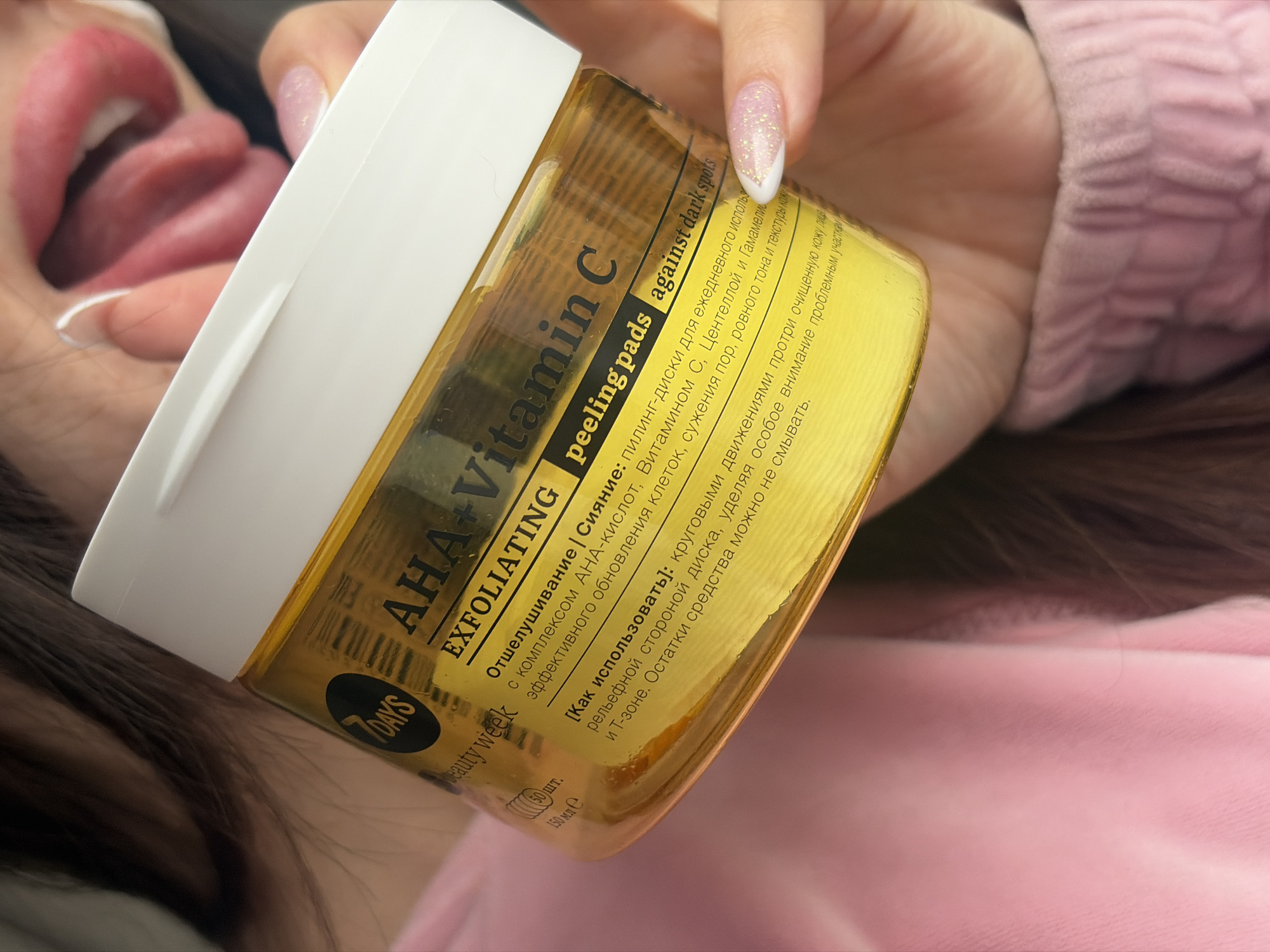 A person is holding a container of "7DAYS" brand "AHA+Vitamin C EXFOLIATING peeling pads" in front of their face, partially obscuring their features. The product is described as "peeling pads against dark spots" and includes instructions in Russian for use on the face. The container appears to be yellow and white. The person's nails are manicured with a white and glitter design. A portion of the person's open mouth and tongue are visible behind the product. They are wearing a pink, textured garment. The location is not specified, and there is no indication that this image was taken in the city of Saint Petersburg, Russia.