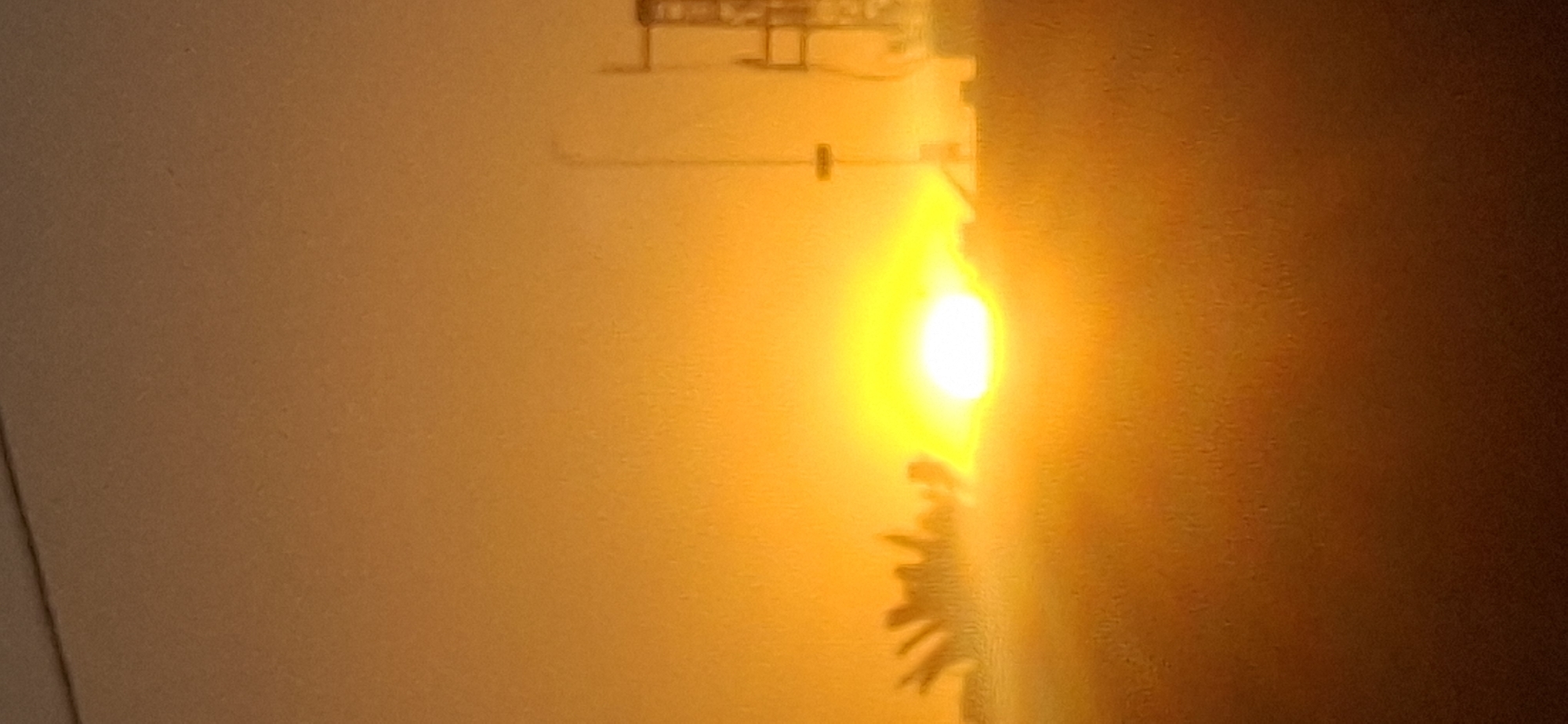 The image captures a sunset in Yerocham, Israel, characterized by a vibrant orange and yellow gradient sky. In the foreground, the silhouette of some vegetation or trees is visible on the left. To the right of the frame, a tall, slender structure, possibly a utility pole or a radio tower, stands against the bright sky. In the far right, part of a larger, industrial-looking structure, likely a tower or building, is also silhouetted. The overall scene is bathed in the warm, diffused light of the setting sun, creating a hazy and atmospheric effect.