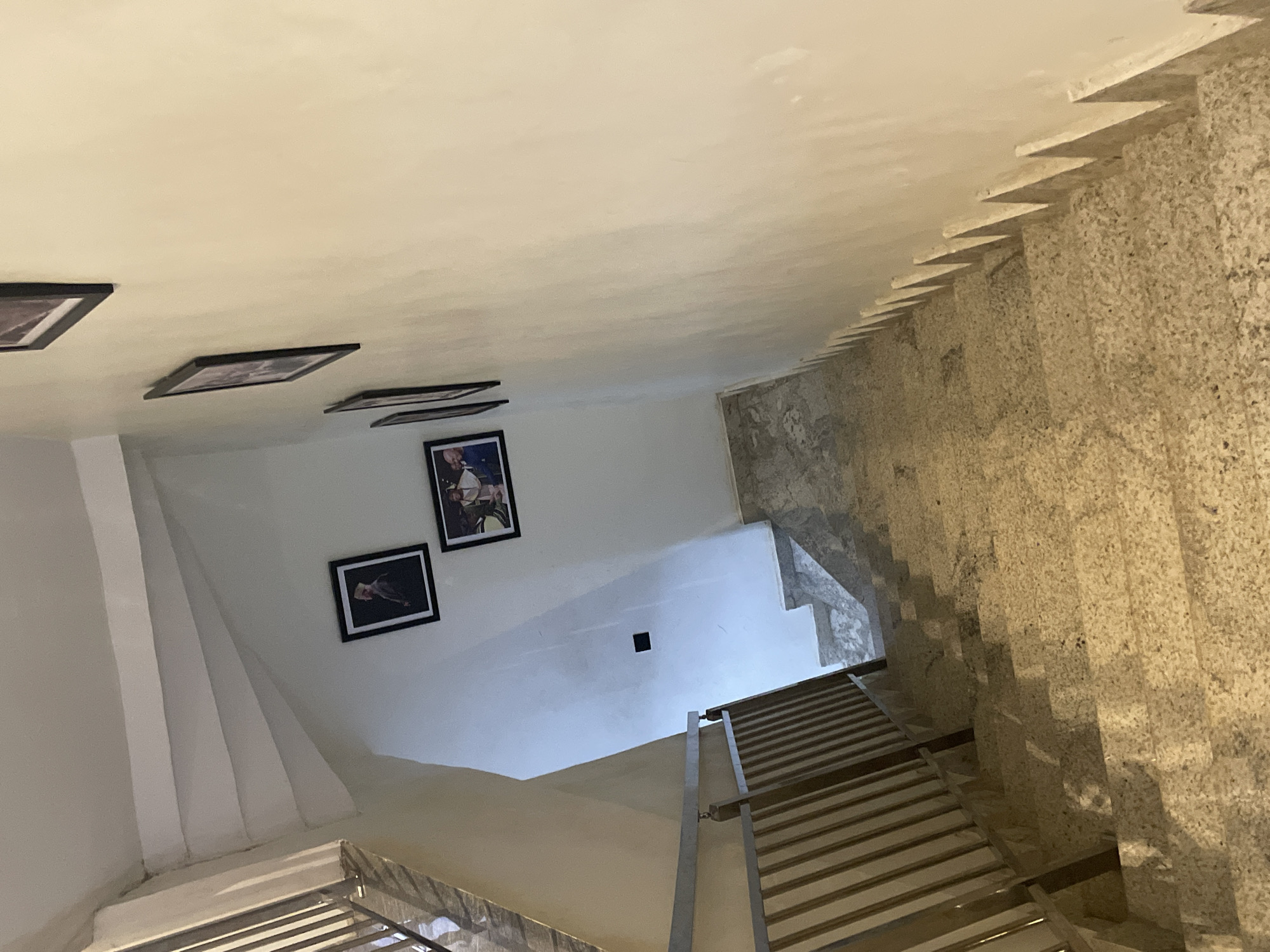 An indoor shot of a well-lit staircase in a building in Abuja, Nigeria. The staircase is made of speckled granite and is bordered by a wooden railing and a white wall. Several framed photographs are mounted on the wall, depicting individuals in formal attire. A dark square electrical outlet is visible on the wall below the photographs. The scene conveys a sense of domesticity and personal history within a modern architectural setting.