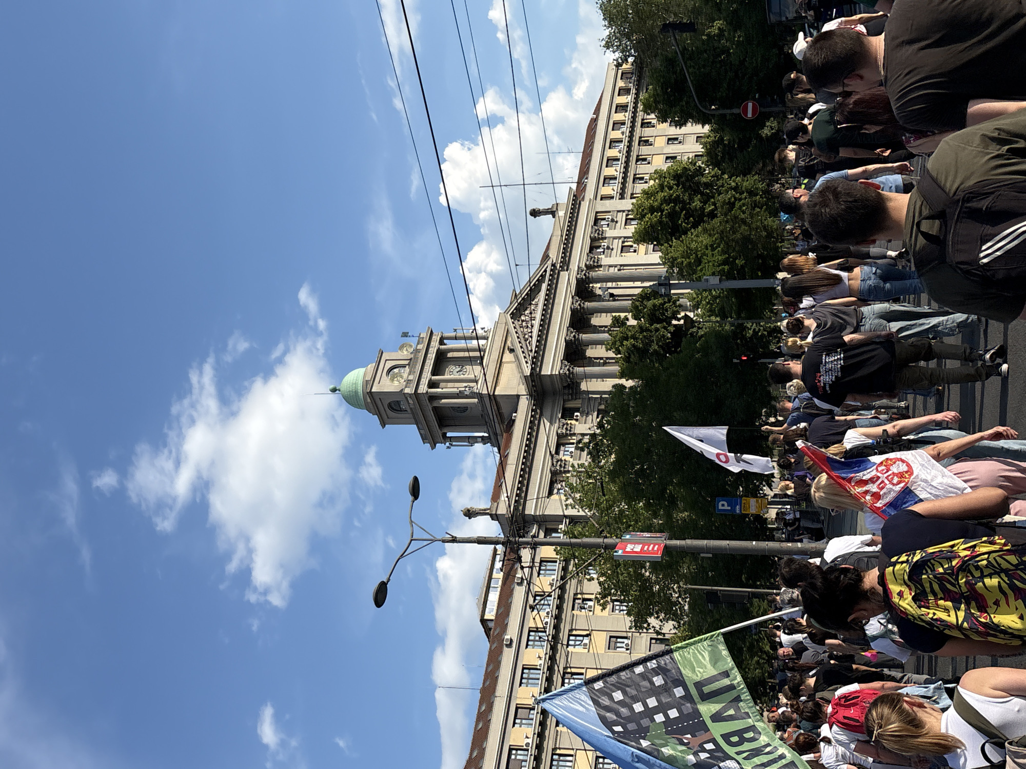 The image shows a daytime street scene in Belgrade, Serbia, possibly during a protest or demonstration. The Serbian Parliament Building is visible in the background, identified by its distinctive clock tower and dome. In the foreground, a crowd of people is gathered, holding various flags and banners. The flags include the Serbian flag, as well as what appear to be protest banners. The scene is set on a sunny day with a blue sky and some clouds. Power lines cross the sky, adding to the urban context. People are densely packed, with the camera positioned in the crowd, looking toward the Parliament Building. A street light is visible in the midground.