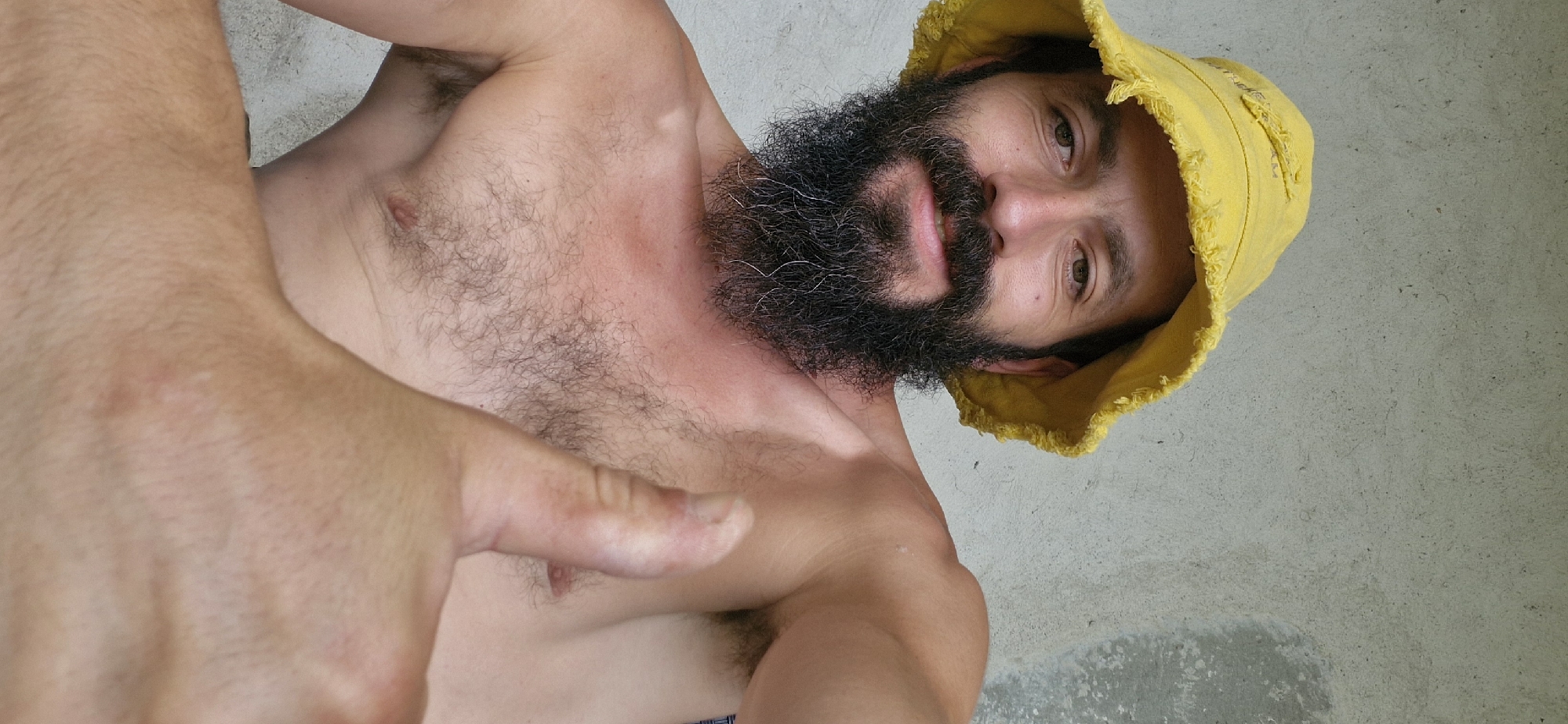 A man wearing a yellow bucket hat and a dark beard is seen in a close-up shot, giving a thumbs-up gesture. He is shirtless, revealing a hairy chest and shoulder. The background is a textured, light-colored wall. The image appears to be a selfie, taken from a low angle.