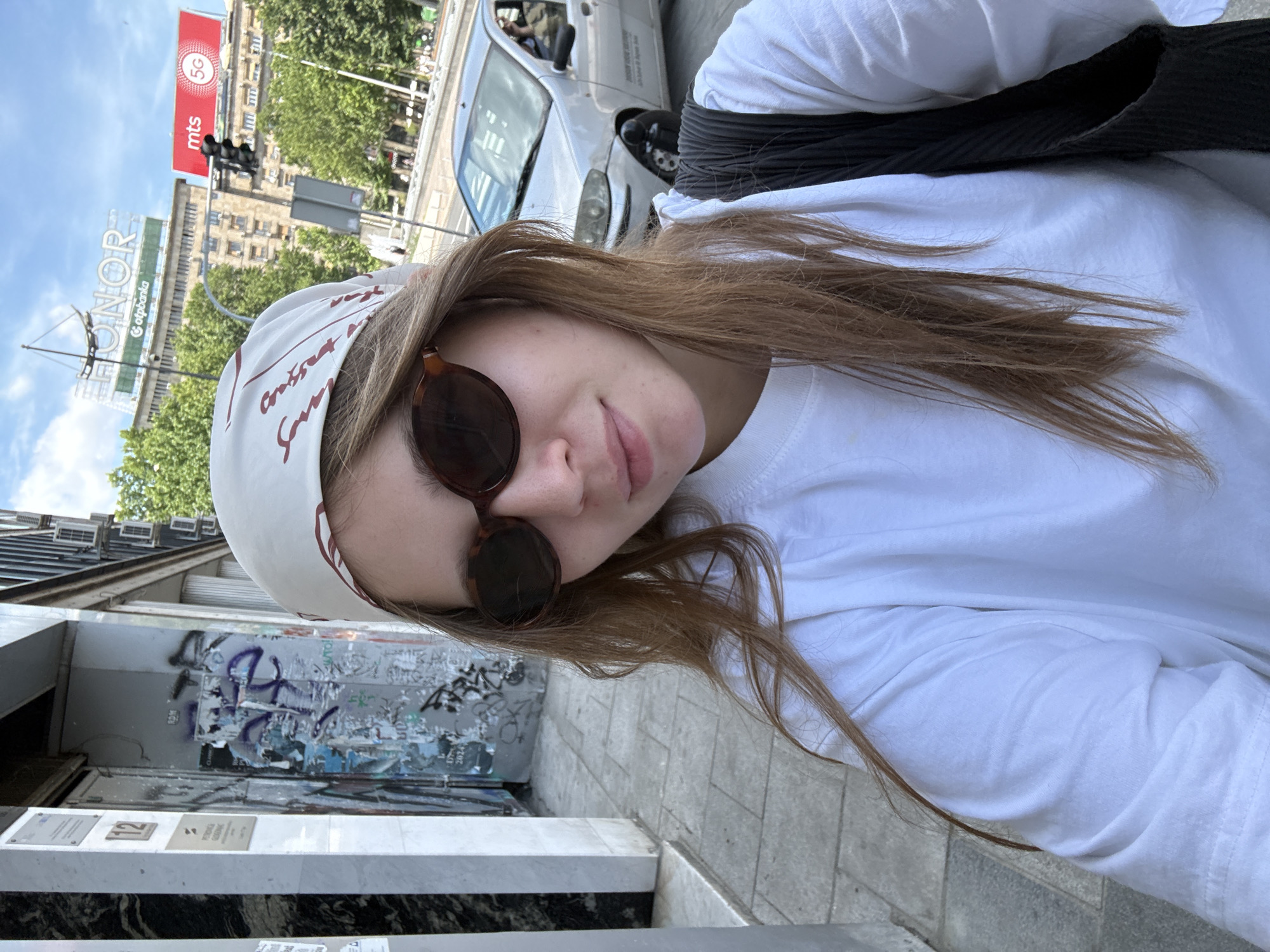 A young woman with long brown hair, wearing sunglasses and a white bandana with a pattern, takes a selfie outdoors in Belgrade, Serbia. She is wearing a white t-shirt and has a black bag on her shoulder. Behind her, a silver car is parked on the street, and a traffic light is visible. In the background, buildings and trees can be seen under a cloudy sky. The woman is in the foreground, with a blurred background that suggests she is standing on a sidewalk in a busy area of the city. The image appears to be taken during the day.