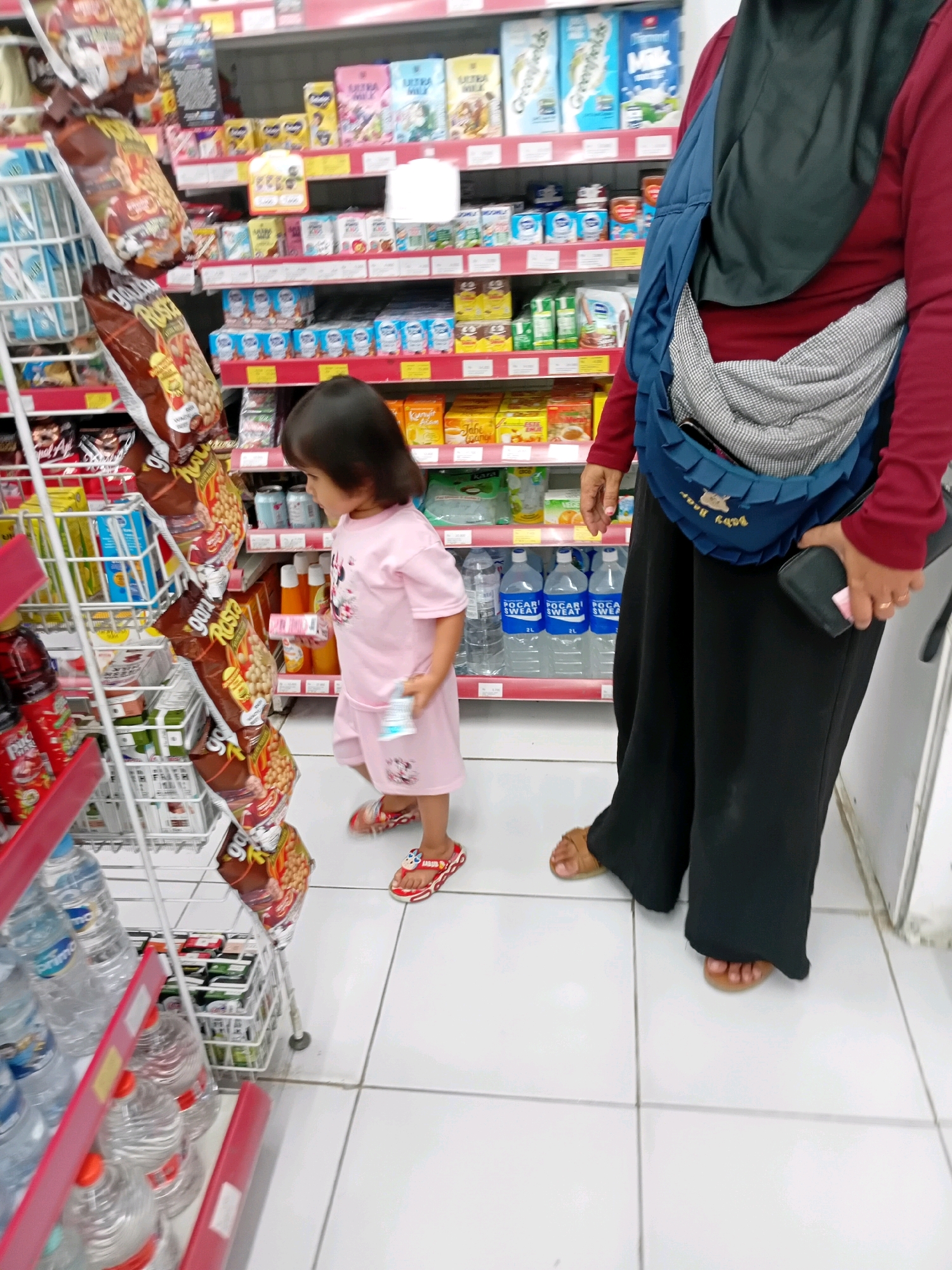 In a brightly lit supermarket aisle in Seren, Indonesia, a young child, likely a girl, wearing a pink t-shirt and shorts with cartoon character prints, stands looking at snacks displayed on a metal rack. She is wearing red flip-flops with a cartoon character. Beside her, a person, likely an adult, wearing a dark hijab, a maroon long-sleeved shirt, and loose black pants, is holding a dark wallet in their right hand and a pink object in their left. The adult is also wearing brown sandals. The shelves are stocked with various food and beverage items, including bottles of "POCARI SWEAT" water. The floor is tiled with white squares. The overall scene depicts a routine shopping trip.