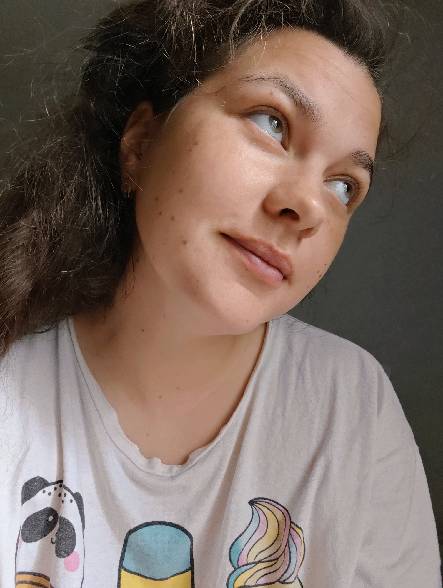 An indoor portrait captures a young woman from Chaikovsky, Russia, in a close-up, angled shot. She is looking upwards and slightly to the right with a contemplative and relaxed expression, her lips hinting at a soft smile. Her long, dark, wavy hair cascades around her shoulders, with some lighter strands visible due to natural light.
The woman has fair skin with scattered freckles and moles on her face and neck. Her eyes are light-colored, reflecting the ambient light. She is wearing a casual, light-colored (likely white or off-white) t-shirt. The shirt features cartoon-style illustrations: a black and white panda head with pink cheeks is visible on the left side, and a colorful swirled design, possibly an ice cream cone with yellow, blue, and pink hues, is on the right side.
The scene is lit by natural daylight, casting subtle shadows and highlighting her features and hair, suggesting a daytime setting. The background is a plain, dark gray wall, providing a neutral backdrop that allows the subject to stand out. No text is visible within the image.