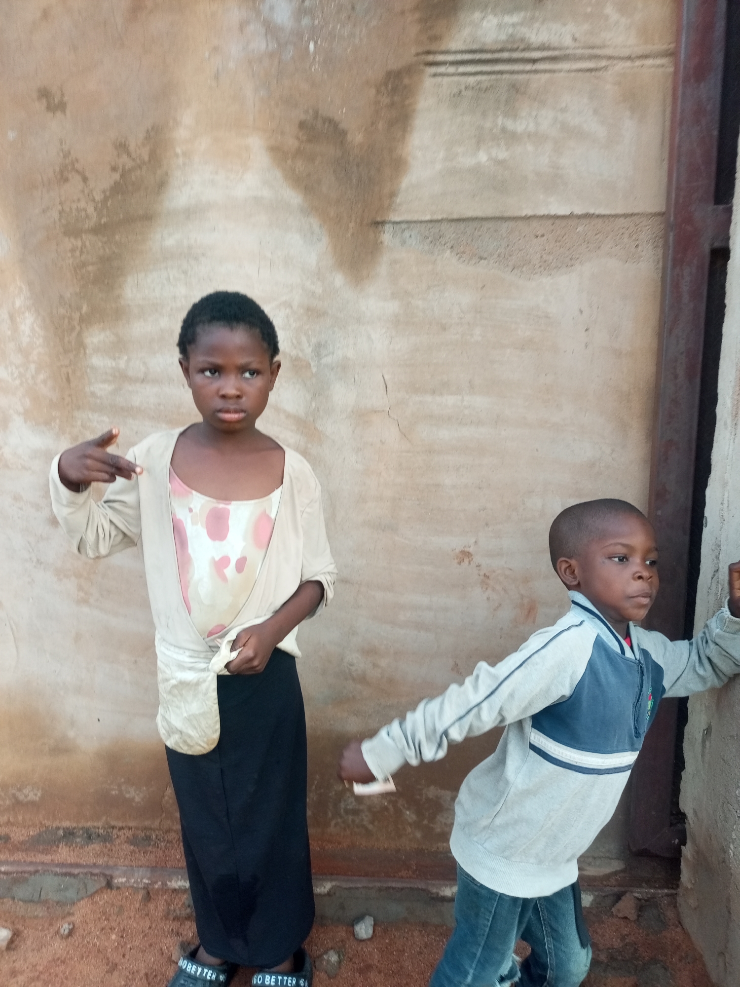 In the city of Tempure, Nigeria, two children stand near a building. The child on the left, a girl, faces the camera and holds her right hand up, making a gesture with her fingers. She wears a patterned dress and a light-colored cardigan tied at the waist, over a dark skirt and black sandals. The child on the right, a boy, is leaning against the building, facing away from the camera. He wears a gray and blue zippered jacket and blue jeans. The background consists of a textured, light-colored wall and a vertical metal post. The ground is covered with reddish sand and small stones.