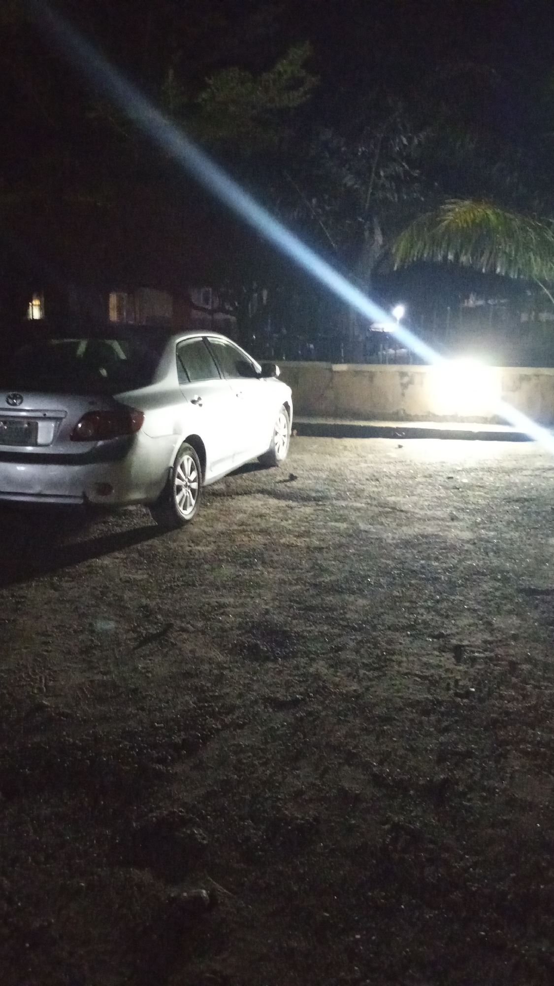 A nighttime outdoor scene is depicted in Adankolo, Nigeria. A silver four-door sedan, consistent with a Toyota Corolla model, is parked on an unpaved, dark ground. The vehicle is angled with its rear-left side towards the viewer, exhibiting a visible dent on its rear-left quarter panel above the wheel arch. The car's tail lights are not illuminated. The ground surface is uneven and appears to be composed of dirt and small gravel. Intense artificial light sources, originating from the mid-background and right, illuminate the area, casting bright light onto a low concrete wall situated behind the parked vehicle. A prominent, diagonal beam of light traverses the upper-mid section of the frame. Dark foliage, including identifiable palm fronds, is present behind the concrete wall and light sources. Faint illumination from distant structures is discernible in the far-left background. No individuals or active interactions are observed within the frame.