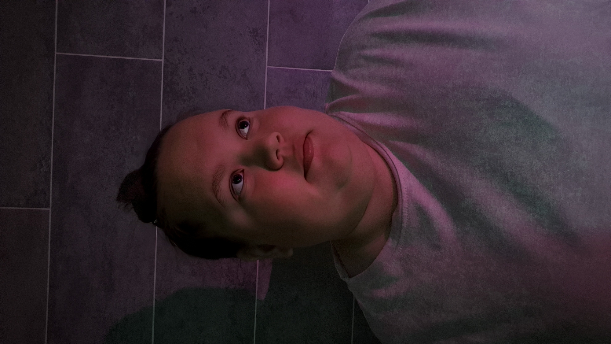 An upward-angled shot captures a person, likely female, with a round face and dark hair pulled back, appearing to lie down or lean back against a tiled surface. The tiles are dark gray with lighter grout lines, suggesting an indoor setting.
The person is wearing a light-colored, possibly gray, t-shirt or hooded top. Their gaze is directed upwards, with a neutral, slightly contemplative expression. The most striking detail is the artificial, colored lighting that bathes the scene. Strong pink and purple hues illuminate the person's face and the front of their shirt, particularly on the right side of their face, giving their skin a reddish glow. Greenish light is also visible, casting a subtle tint on the left side of their face and shirt, blending into the purple tones on the tiled background. The lighting creates a dramatic and atmospheric mood.
There are no visible texts or specific location cues within the image itself beyond the tiled surface. The overall impression is one of introspection or a moment captured under unusual, stylized lighting conditions, likely in Chaikovsky, Russia.