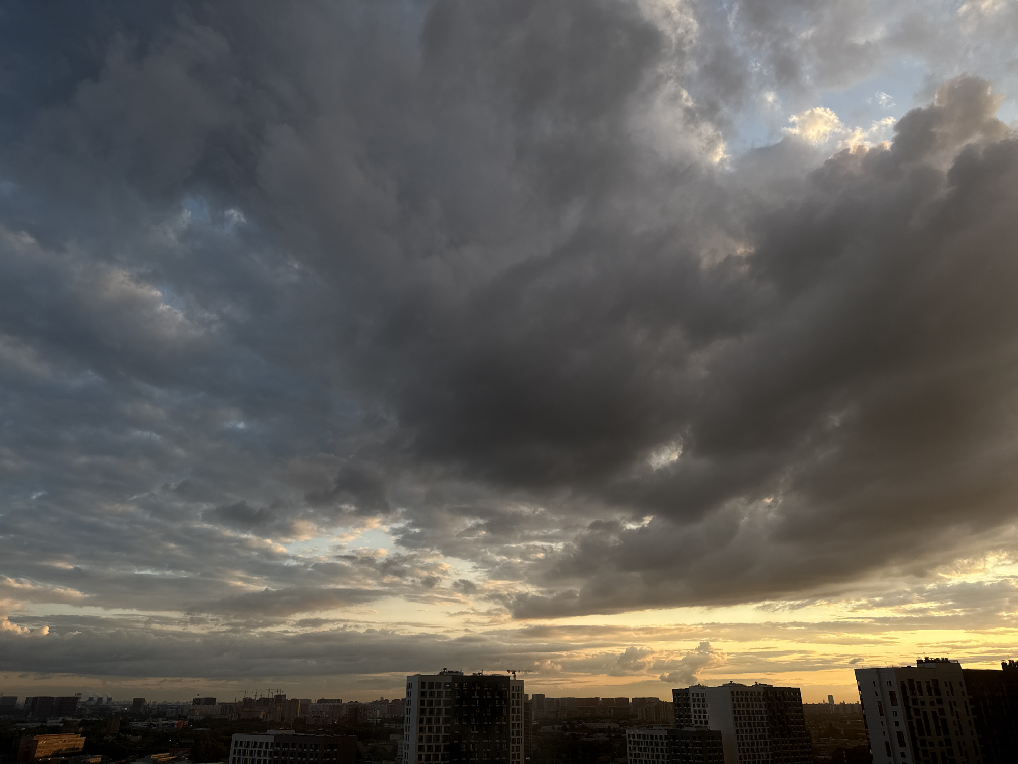 A city skyline under a dramatic sky is captured from an elevated perspective, likely a high-rise building in Tomilino, Russia. The foreground is dominated by dark, heavy storm clouds, interspersed with patches of lighter sky and rays of sunlight breaking through. Below the clouds, the mid-ground features a dense arrangement of modern apartment buildings, some appearing under construction with cranes visible. The buildings vary in height and architectural style, with several taller, more contemporary structures dominating the view. In the background, the cityscape extends further, with more buildings and hints of infrastructure visible. The lighting suggests it is either early morning or late afternoon, with the sun casting a warm glow on the horizon and illuminating the undersides of some clouds. No people are discernible in the image.