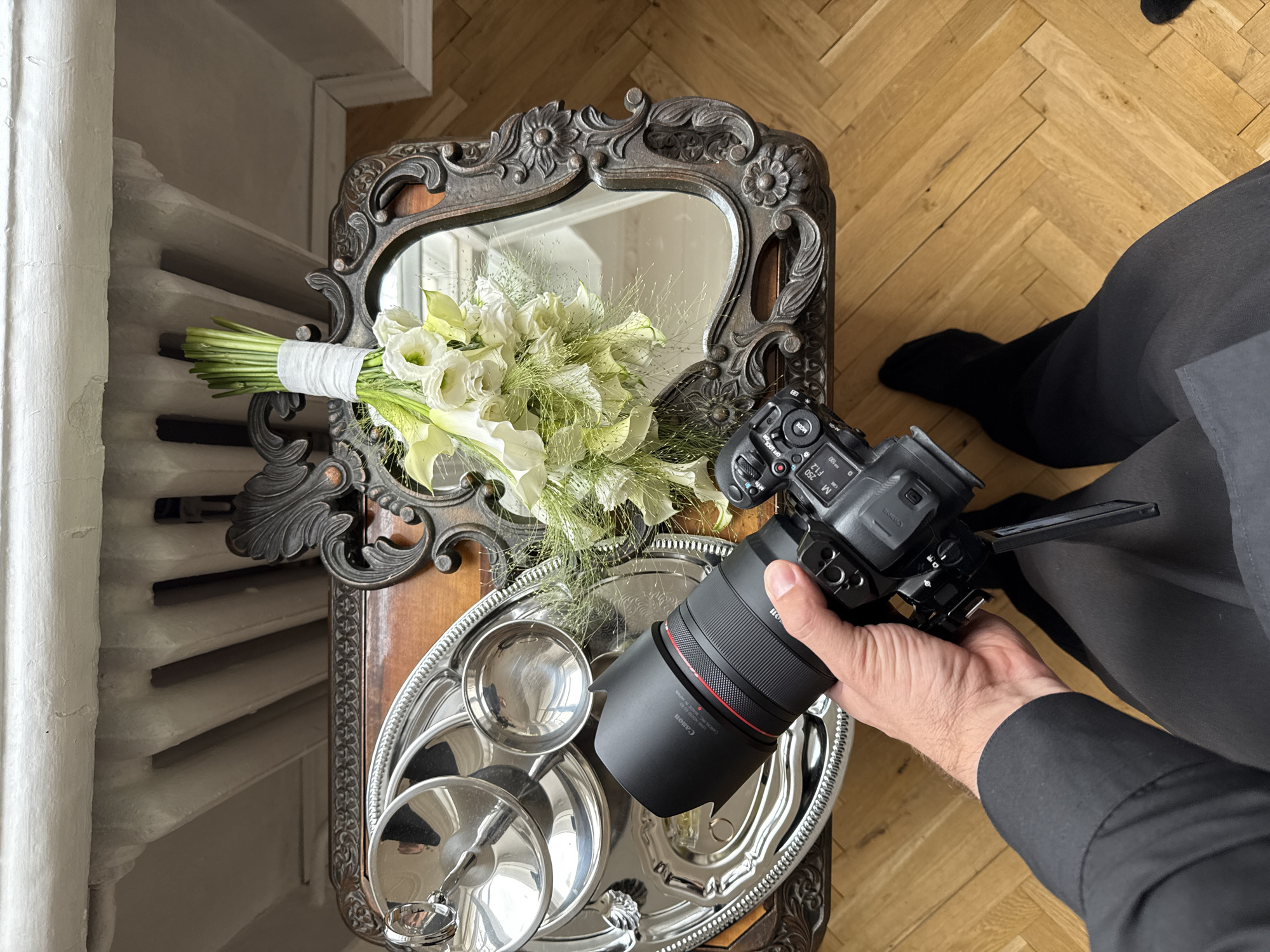 A high-angle view captures a person's right hand holding a black Canon mirrorless camera with an attached black RF lens featuring a red ring and lens hood, displaying camera settings M, 250, F1.2, ISO 100. The person wears a black long-sleeved garment and dark trousers, with black socks on a light brown herringbone parquet floor. The camera is directed downward towards an ornate wooden table situated in Perm, Russia. On the table's left, a decorative silver tray holds a lidded silver serving dish and two small silver cups, reflecting parts of the room. On the table's right, a dark, ornately carved framed mirror is positioned. Resting on the mirror and table is a bridal bouquet composed of white calla lilies and other white flowers, accented with green foliage, its stems bound with white fabric. The background features a white-painted wall with a visible cast-iron radiator underneath. The paint on the wall exhibits minor wear.
