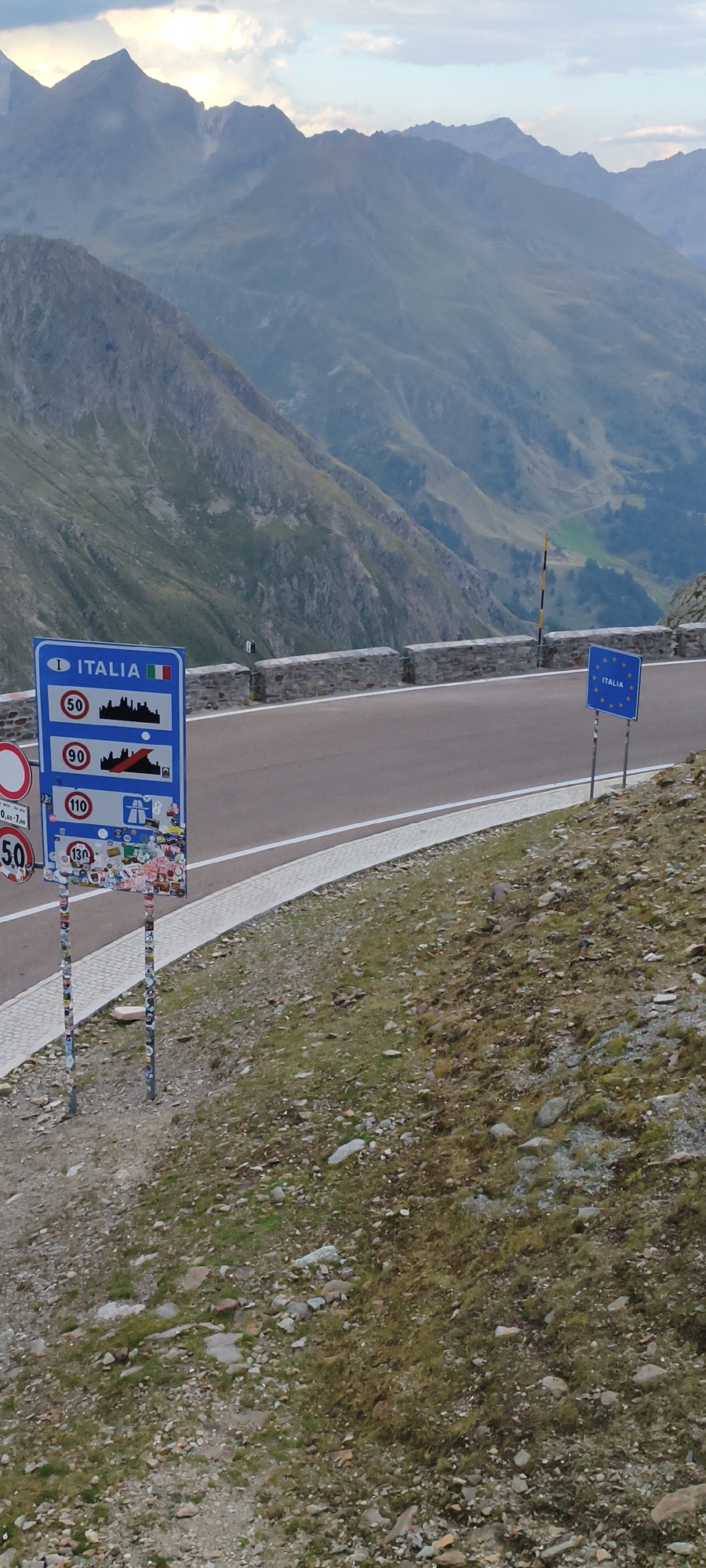 This image depicts a mountainous landscape with a road and signage, likely at a border crossing. The foreground shows a grassy, rocky slope. A road curves into the distance, bordered by a stone wall on the left. Several road signs are visible. The most prominent is a blue sign with white text reading "ITALIA" and the Italian flag, accompanied by circular speed limit signs showing 50, 90, and 110, as well as other symbols. To the right, near the road, a blue European Union sign with stars is posted. The background features a vast mountain range with steep slopes and peaks under a cloudy sky. The location is identified as the city of Sölden, Austria.