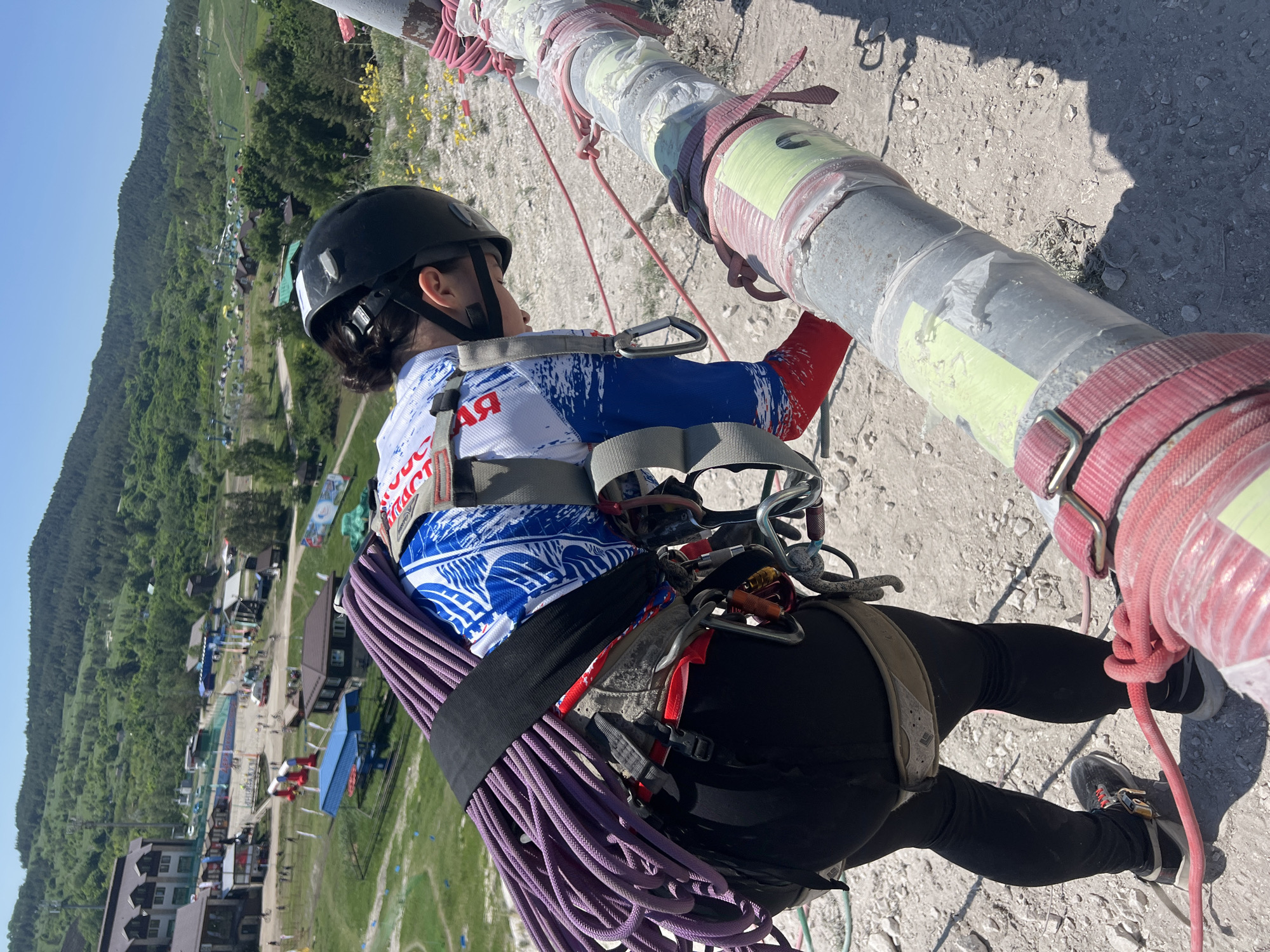 A woman wearing a black helmet, a white, blue and red shirt, black pants, and climbing gear is suspended in the air next to a grey metal pipe. She is wearing a harness and is attached to the pipe with ropes and carabiners. She has a coiled purple rope attached to her harness.
The background shows a green landscape with trees and buildings. It is located in the city of Khvalynsk, Russia.