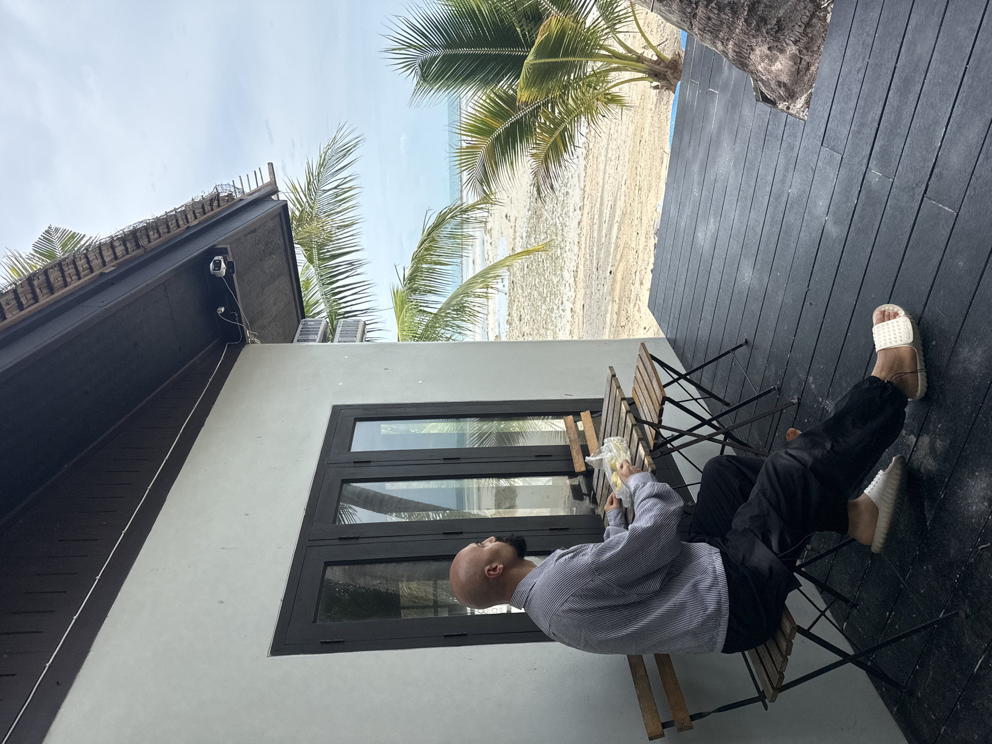 A bald, bearded man sits at a small wooden table on a dark wooden deck, facing left. He wears a vertically striped long-sleeved shirt and black pants, and white slides on his feet. The man holds a clear plastic bag containing yellow objects. Two small wooden chairs and a table are positioned on the deck. Behind the man, a building wall with dark-framed windows and doors is visible. Beyond the deck, a sandy beach meets the ocean under a cloudy sky. Palm trees are present on the beach and near the deck. The scene is located on Ko Samui District, Thailand.
