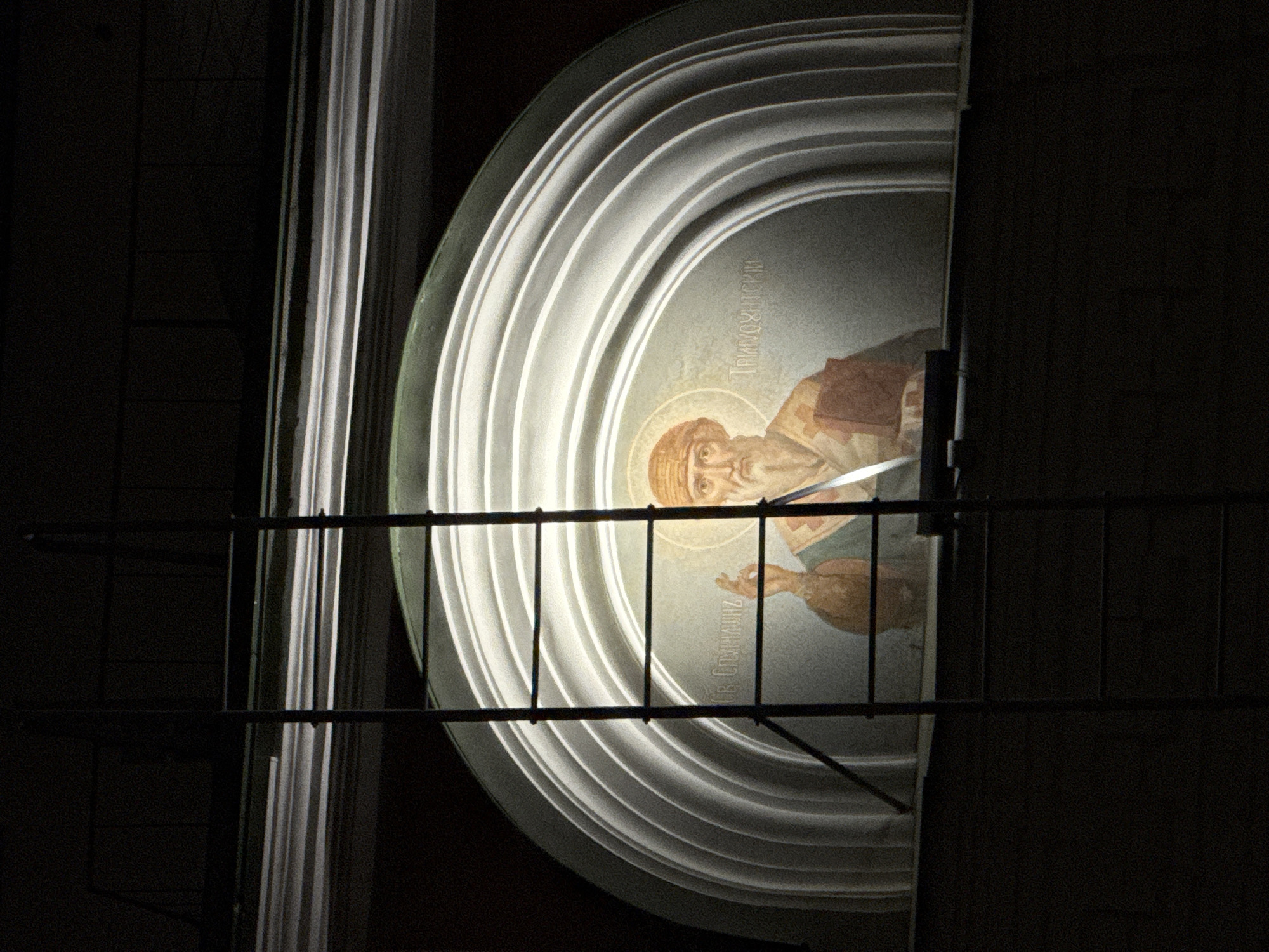 The image depicts an illuminated icon of a religious figure, possibly Saint Spiridon, encased within a rounded architectural element with concentric rings. The icon is located above a doorway or entrance. A metal ladder obscures part of the view. The image appears to be taken at night, and the surroundings are dark. The inscription in the icon is in Russian, which suggests a possible location in Russia.