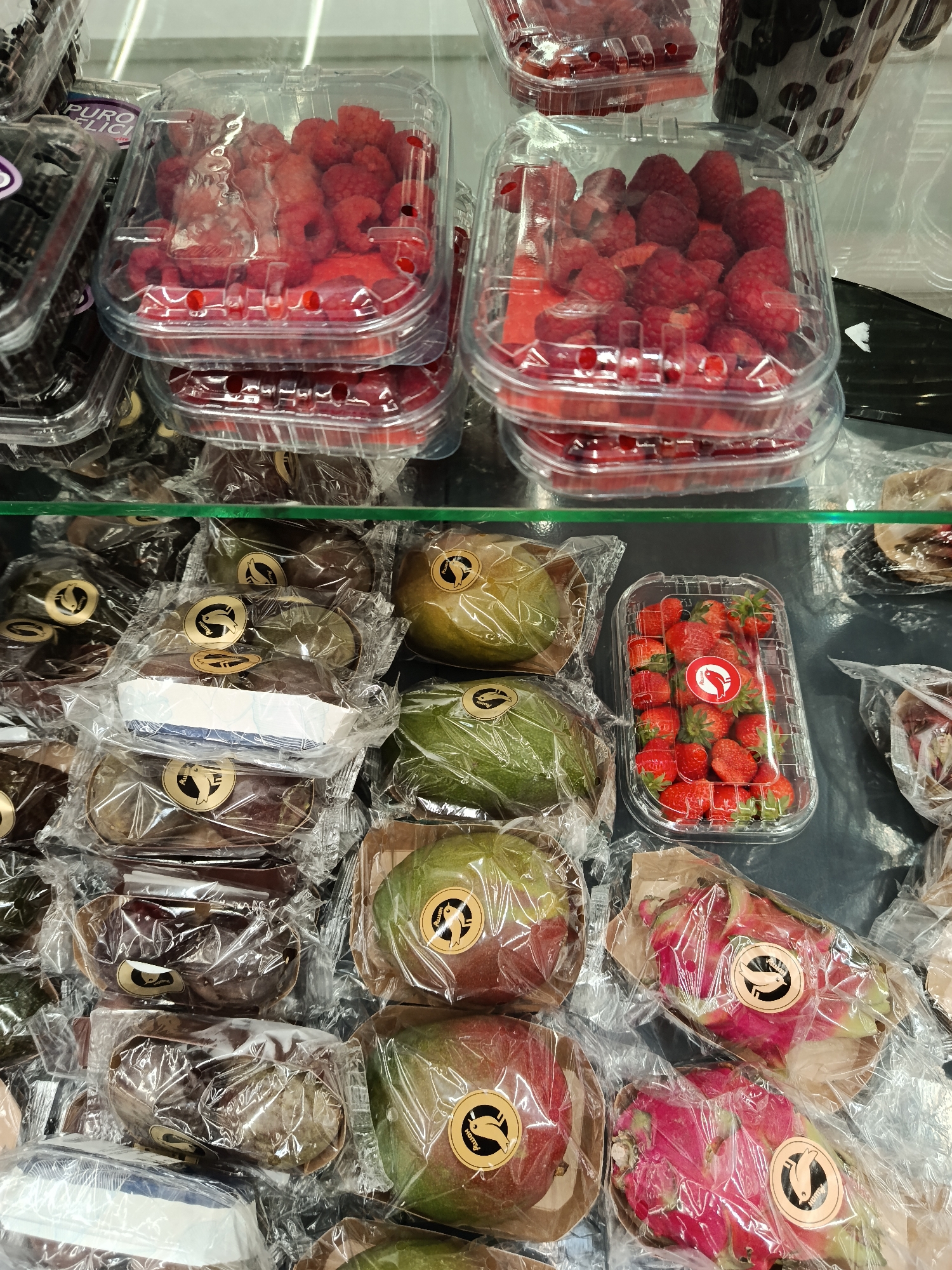 The image shows a refrigerated display case in a store, filled with various packaged fruits. There are several clear plastic containers of raspberries stacked on top, as well as a container of strawberries. Below the berries are items wrapped in plastic, including mangoes, chocolate cakes in paper boats, and dragon fruit. Each individual item has a sticker with a graphic of a leaping fish. The fruits appear to be for sale in a store with expensive presentation. The location is in Moscow, Russia.