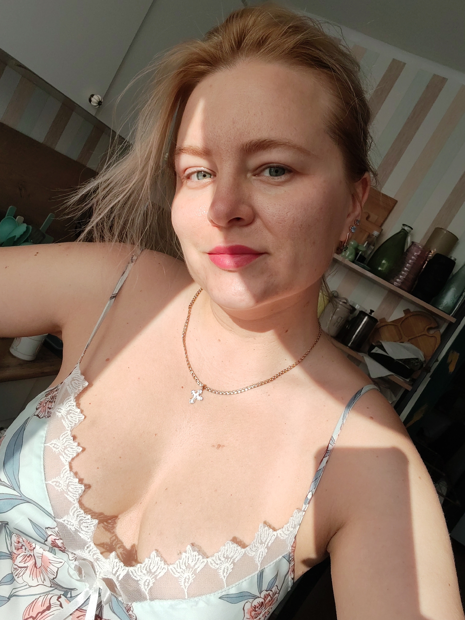 A close-up selfie shows a woman with blonde hair, fair skin, and light blue eyes, looking directly at the camera. She is wearing a light blue silk nightgown with a floral pattern and white lace trim. A delicate gold chain with a crystal cross pendant rests on her décolletage. Sunlight casts a shadow across her face, highlighting one eye and cheek. The background appears to be an interior space with shelves filled with various items, including bottles and containers, against a striped wallpaper. The overall impression is casual and intimate. The image does not contain any information that can be used to determine the location in Izhevsk, Russia.