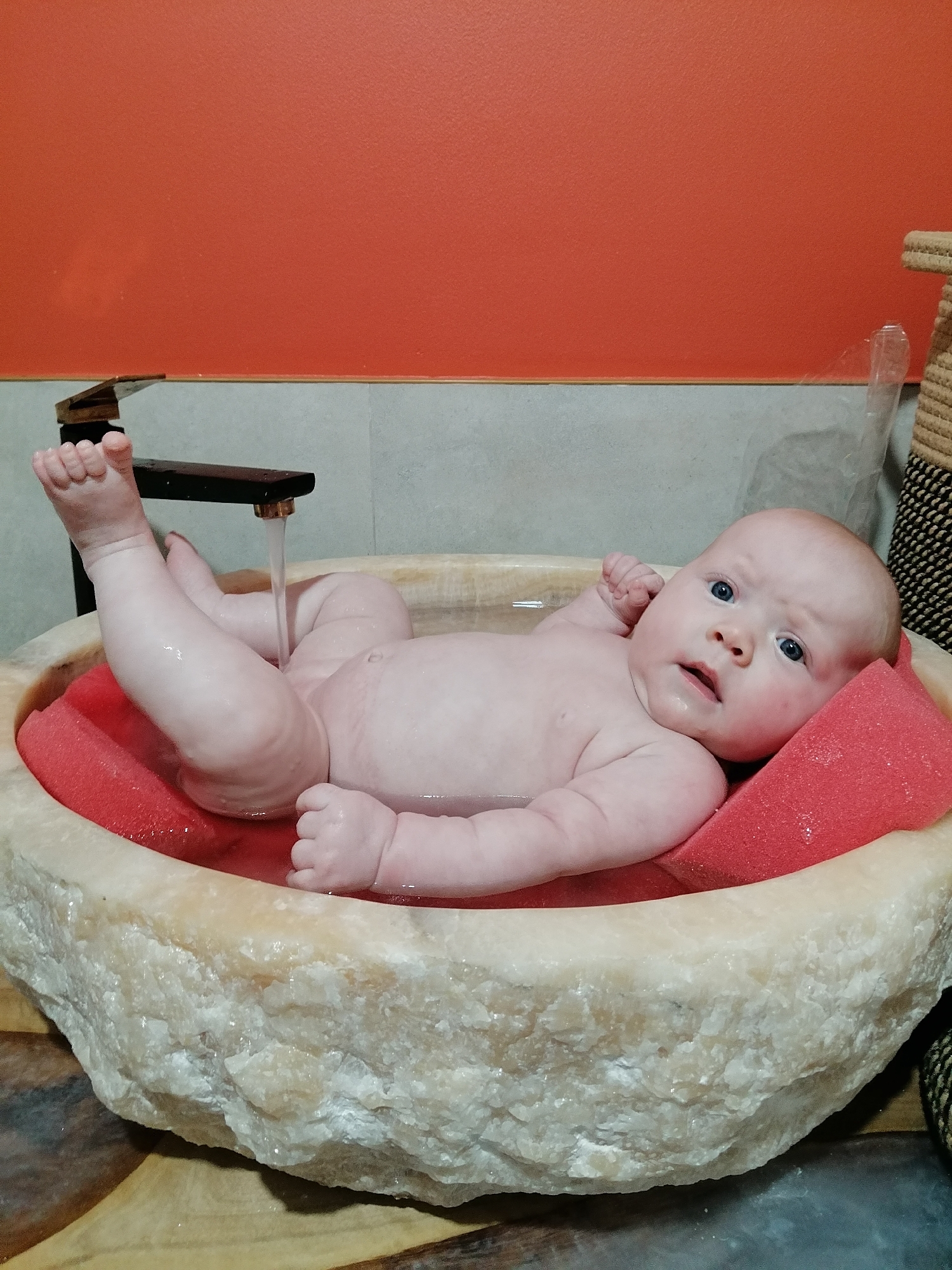 This image captures a baby being bathed in a unique, rough-hewn stone basin. The baby, with wide blue eyes and chubby limbs, lies on a bright red sponge. Water streams from a modern faucet into the basin, creating ripples on the surface and wetting the baby's skin. The background features an orange-painted wall and a textured grey tiled section. To the right, a woven basket is partially visible. The lighting suggests an indoor setting, possibly a bathroom. The baby appears calm and alert, with a slightly surprised expression. There is no visible text in the image. The setting hints at a home environment in Trubacheyevka, Russia.