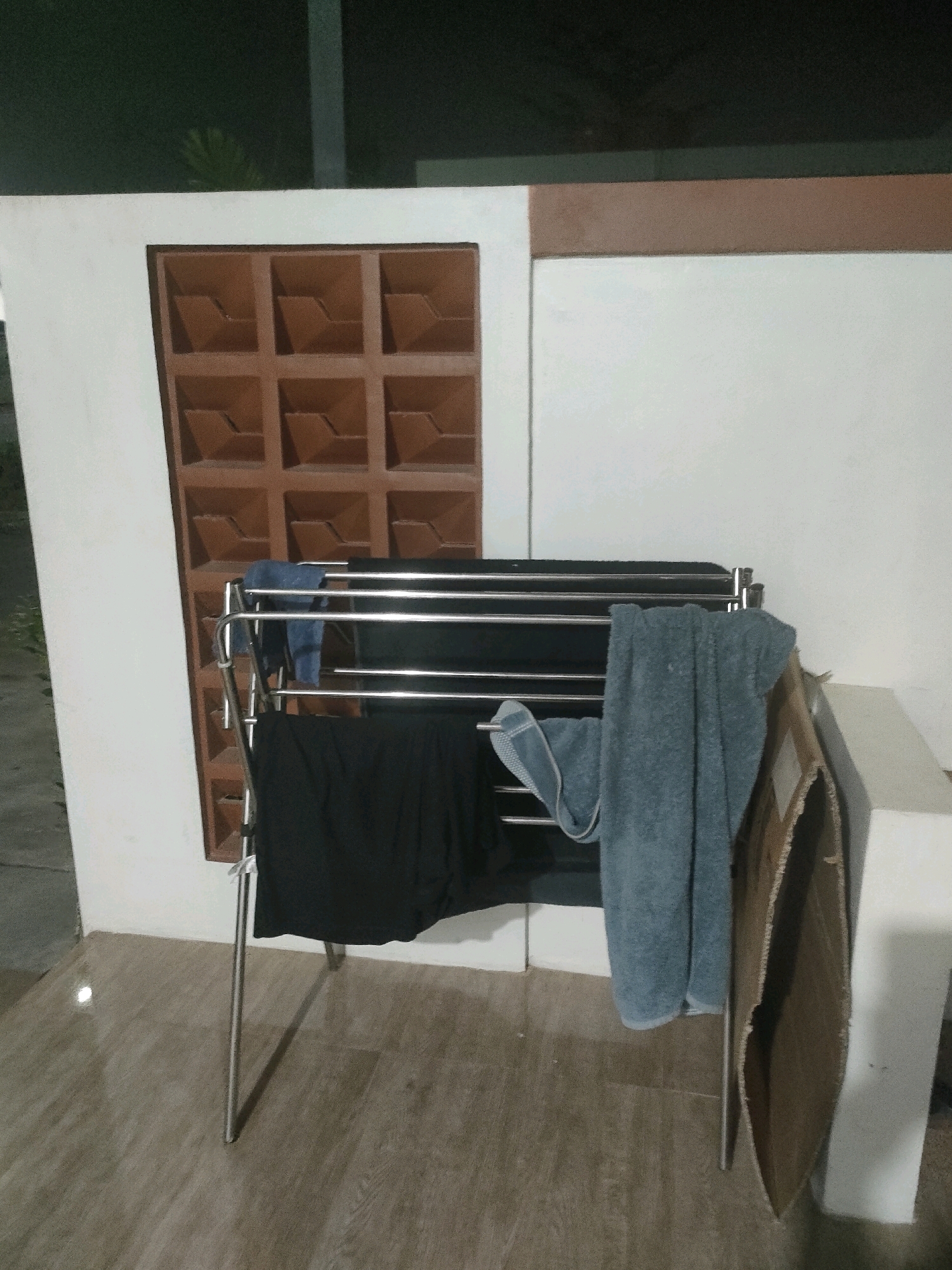An outdoor drying rack with metal poles holds several pieces of clothing, including a black garment and two blue towels. To the left of the drying rack is a wall with a decorative square lattice vent. The rack stands on a wooden-patterned floor. In the background, a dark, out-of-focus area suggests foliage or a nighttime scene. A piece of cardboard is leaning against the wall to the right of the drying rack. The scene appears to be in Batam, Indonesia, likely on a patio or balcony given the outdoor setting and the visible architecture.