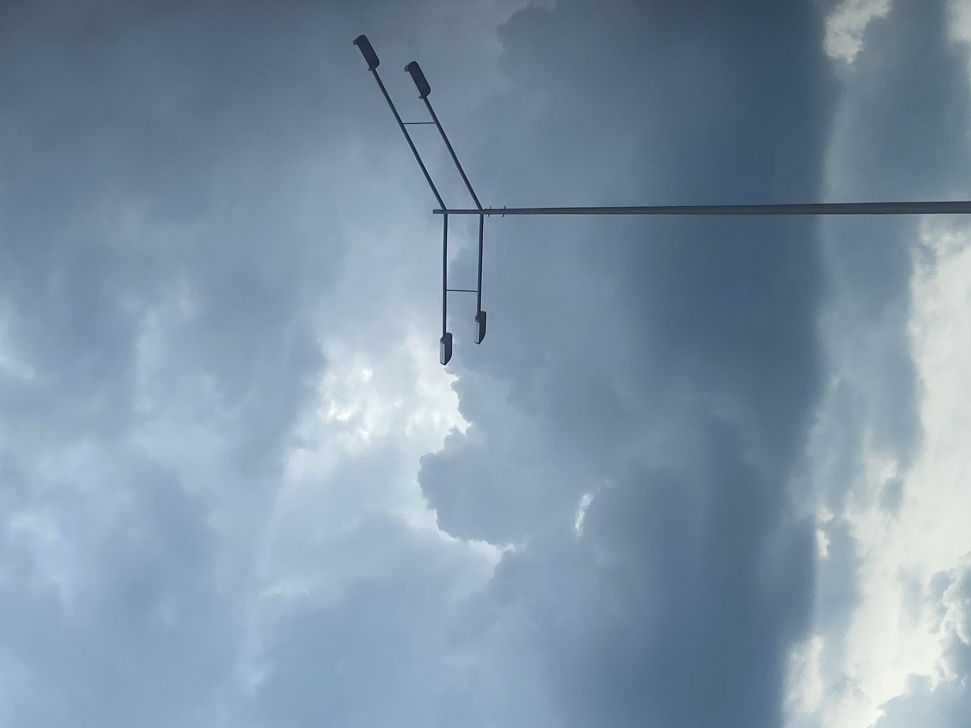 A low-angle shot captures a sky dominated by thick, grey storm clouds. In the upper portion of the frame, a metal utility pole extends horizontally. Attached to the pole is a vertical arm supporting two streetlights. Isakovo, Russia.