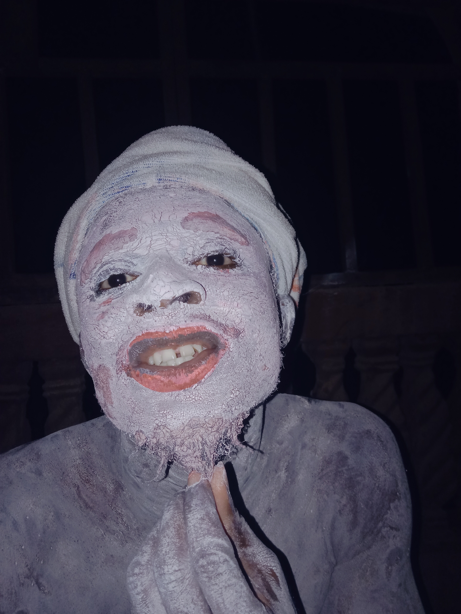 The image presents a headshot and upper body view of a person against a dark, out-of-focus background that suggests a dimly lit environment. The subject's face and visible upper body are covered in a thick, cracked, white substance, resembling dried paint, clay, or chalk. This substance gives their skin a highly textured appearance, with hints of the natural skin tone, possibly reddish, visible through the cracks, especially around the eyes and chin.
The person is wearing a light-colored, towel-like cloth wrapped around their head, forming a turban. Their lips are painted a distinct reddish-orange color, and they display a wide, gapped smile, revealing their white teeth. Their dark eyes are open and look directly towards the camera, conveying an engaging and somewhat jovial expression. One of their hands, also covered in the white substance, is brought up to touch their chin.
The context of the person's appearance, with the distinctive body and face covering, suggests a cultural event, performance, ritual, or artistic expression. The dark background accentuates the bright, transformed figure. There is no visible text in the image.