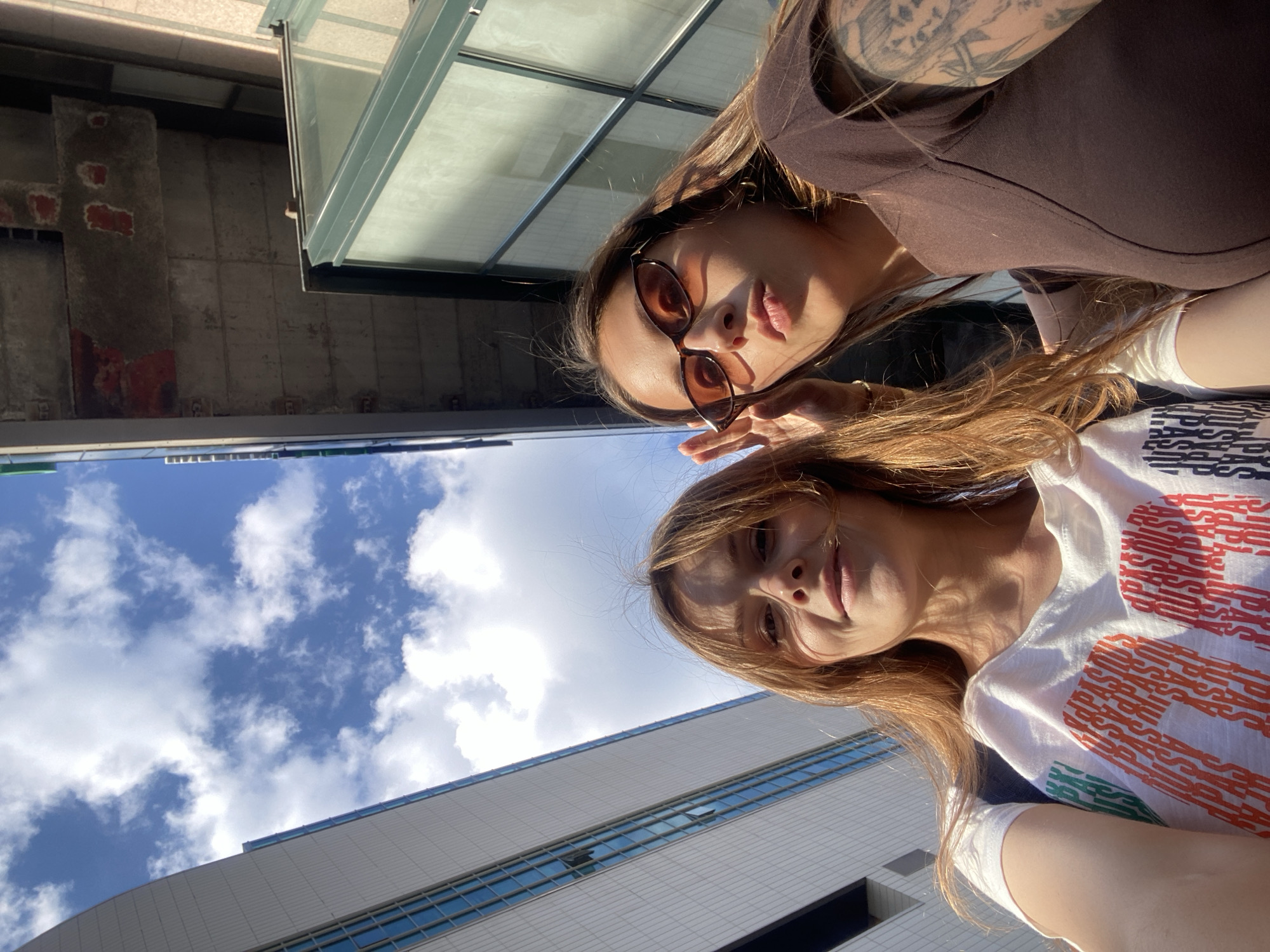 An outdoor, low-angle photograph, taken in Sinyavino, Russia, depicts two adult females in the foreground against a backdrop of urban buildings and a partly cloudy sky. The woman on the left has shoulder-length, reddish-brown hair and fair skin, wearing a white t-shirt adorned with multicolored, repeated text. She smiles subtly towards the camera. The woman on the right has long, dark brown hair, tanned skin, and wears narrow, brown-tinted sunglasses and a dark brown short-sleeved top. Her left arm displays an extensive tattoo sleeve. She holds her right hand near her temple, and her lips are slightly parted in a soft pout. Strong sunlight illuminates both individuals. The background features a blue sky with scattered white cumulus clouds. To the left, a modern, light-colored building with multiple blue-paned windows is visible. To the right and overhead, an older, concrete structure exhibits exposed rebar and patches of red paint, adjacent to a dark green-framed glass architectural element.