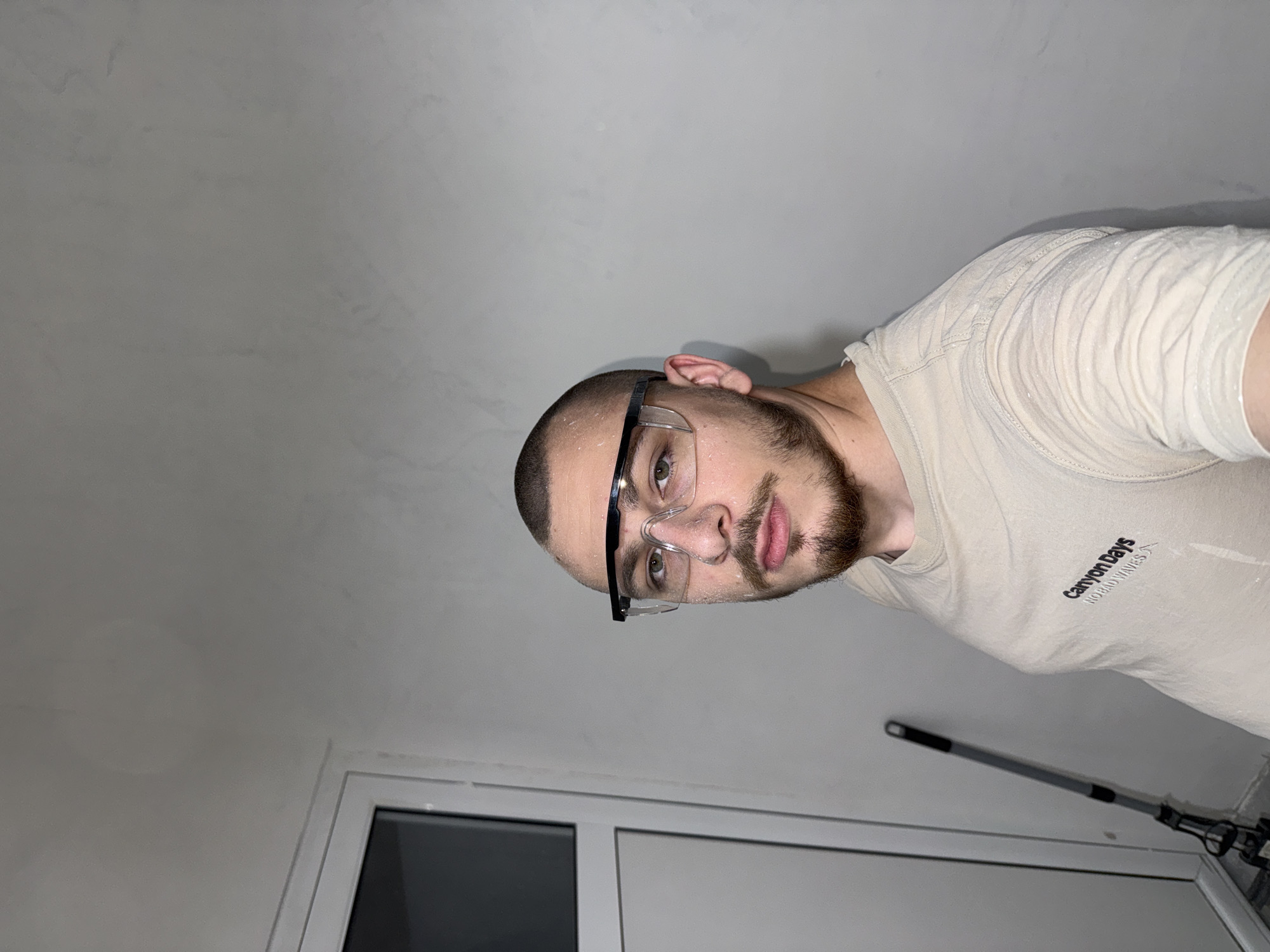 A man with a shaved head and a short beard is wearing safety glasses. He's wearing a light-colored t-shirt with the words "Canyon Days NO BAD WAVES" printed on it. The man is indoors, possibly in a room under renovation, as indicated by the presence of a cleaning tool leaning against the wall. The room has white walls and a window frame is visible. The location is unknown, but the person may live in the city of Stara Zagora, Bulgaria.