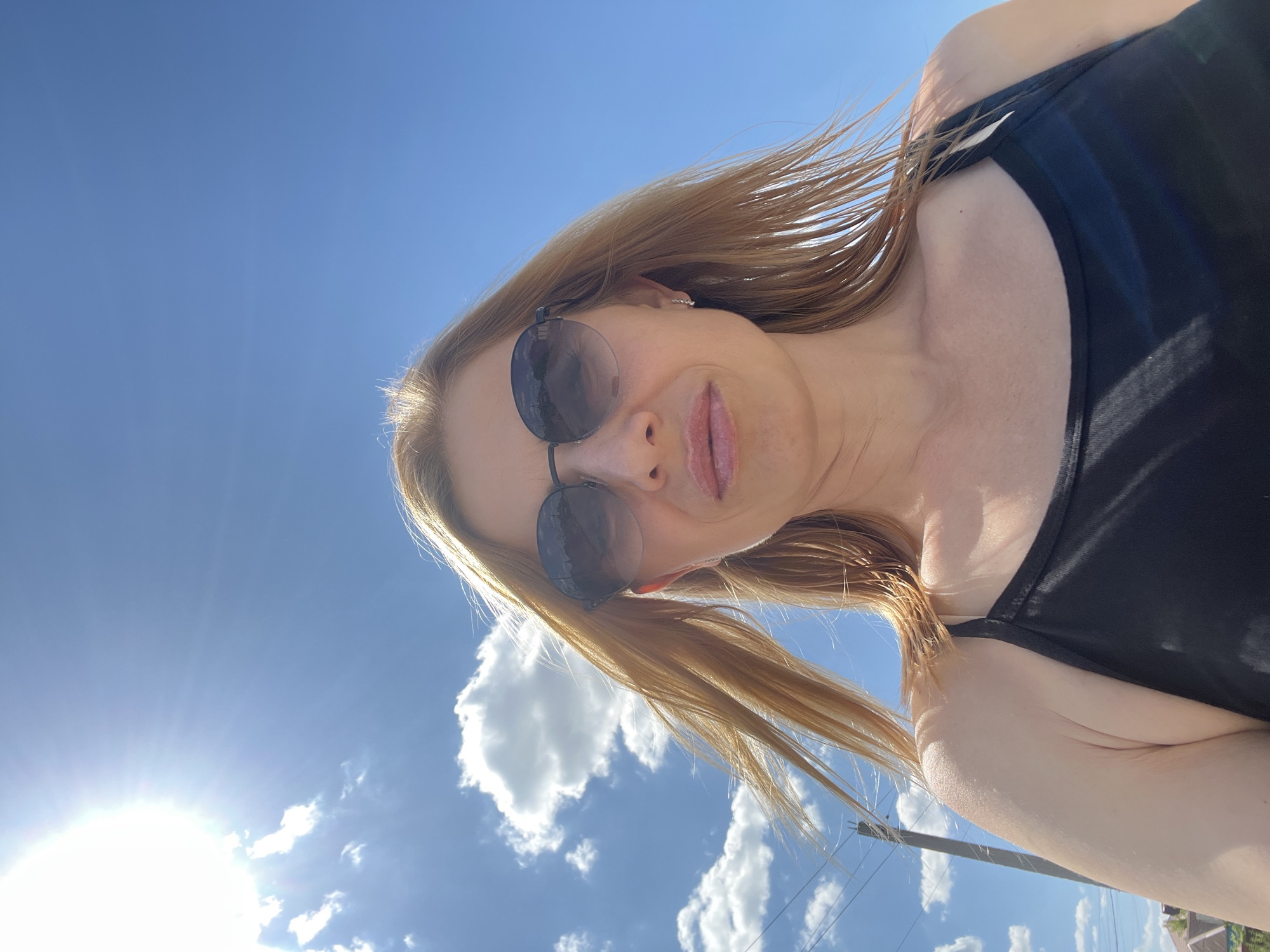 A low-angle, medium shot features a blonde woman in her late 30s or early 40s wearing sunglasses and a black tank top, taking a selfie outdoors under a bright blue sky with scattered white clouds. The sun is high and bright, casting a glare in the upper left corner of the image. The woman's blonde hair is blowing in the wind. She is wearing subtle makeup, with a touch of pink on her lips and small, sparkling earrings. Her expression is neutral, with her lips slightly parted. The background shows a utility pole and some distant houses with red roofs, suggesting a suburban or rural setting. The overall lighting is harsh due to the bright sun. Based on the provided context, the location is identified as Bataysk, Russia.