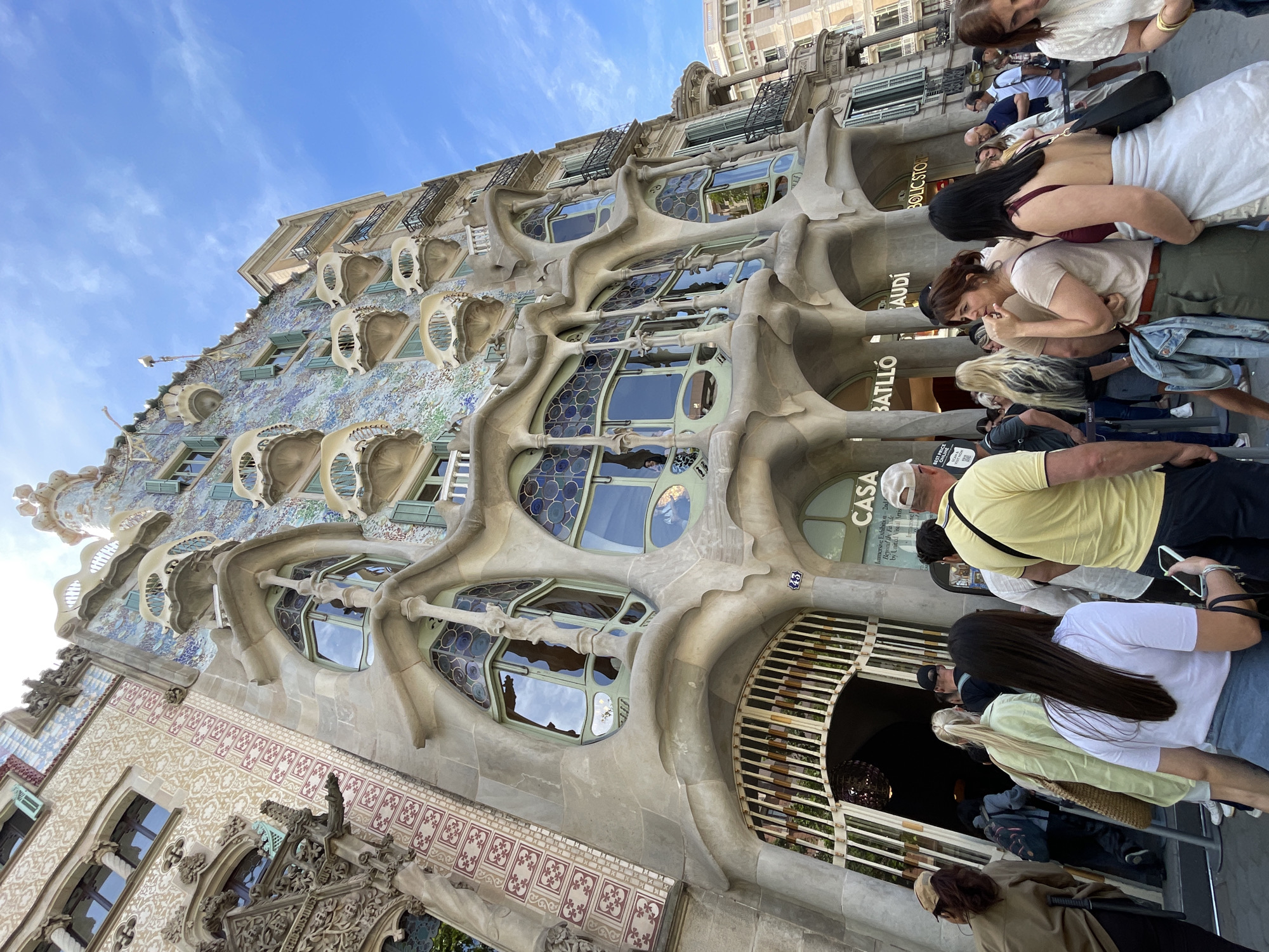 The image depicts Casa Batlló in Barcelona, Spain. The building's unique architectural style, characterized by organic, flowing shapes and colorful mosaics, is prominent. A group of people is gathered near the entrance. Some individuals are looking at the building, while others seem to be waiting in line. The sky is visible in the background, with a clear blue hue. The building's facade features distinctive window designs and sculpted details. The overall scene captures the architectural landmark and its interaction with visitors.