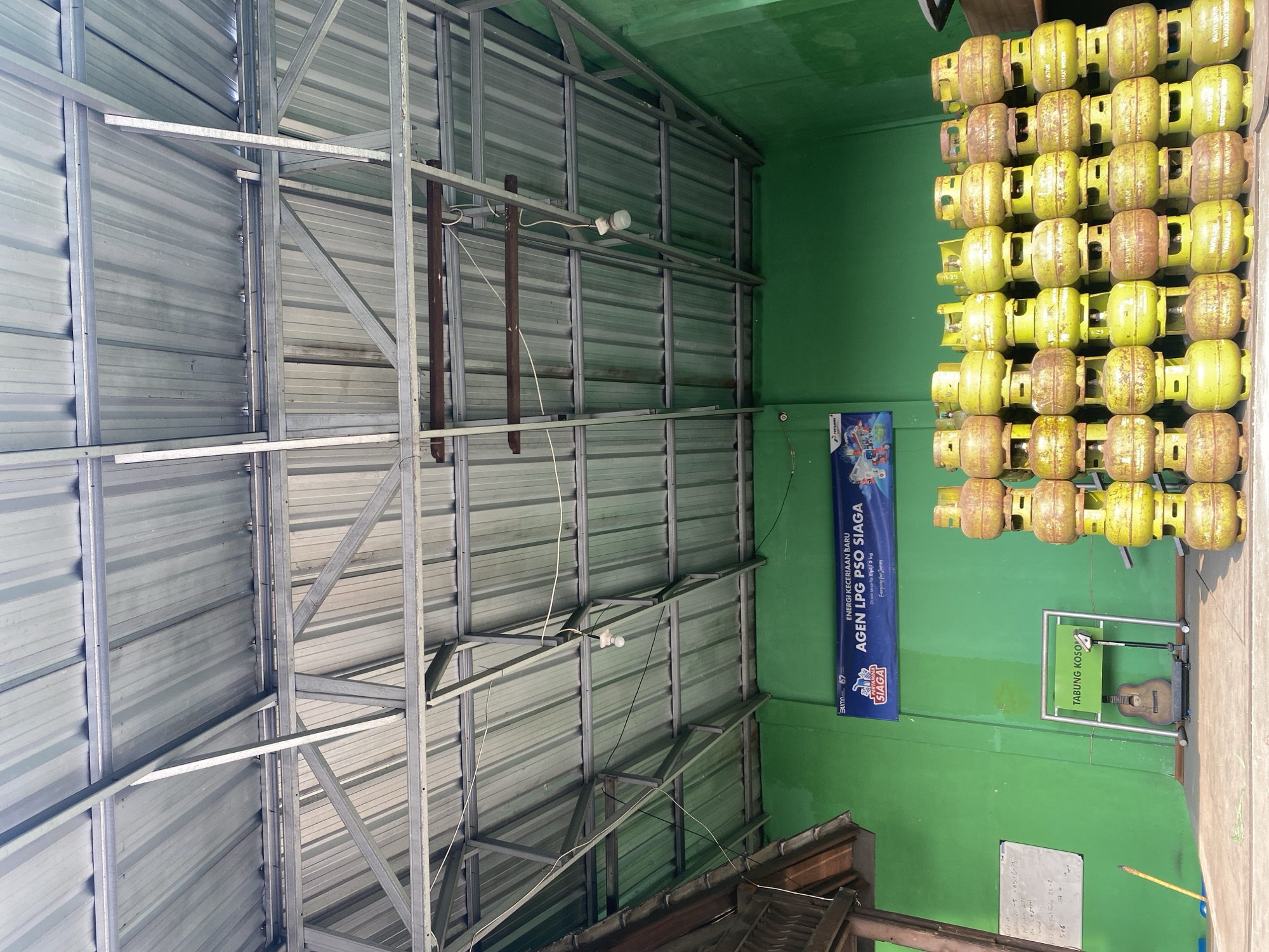 In Tangerang, Indonesia, a storage area features a green wall adorned with a banner for "AGEN LPG PSO SIAGA" (Ready LPG PSO Agent) and the Pertamina logo, indicating a fuel distribution point. The space is filled with stacks of yellow LPG gas cylinders, primarily 3kg size, some showing signs of rust. In the center of the image, a weighing scale is positioned in front of a rack labeled "TABUNG KOSONG" (Empty Cylinder), with an acoustic guitar placed on the scale. A whiteboard with handwritten notes is visible on the left, and a yellow broom leans against the wall. The ceiling is constructed of corrugated metal sheeting supported by a metal frame, with exposed electrical wires and light fixtures. The floor appears to be concrete.