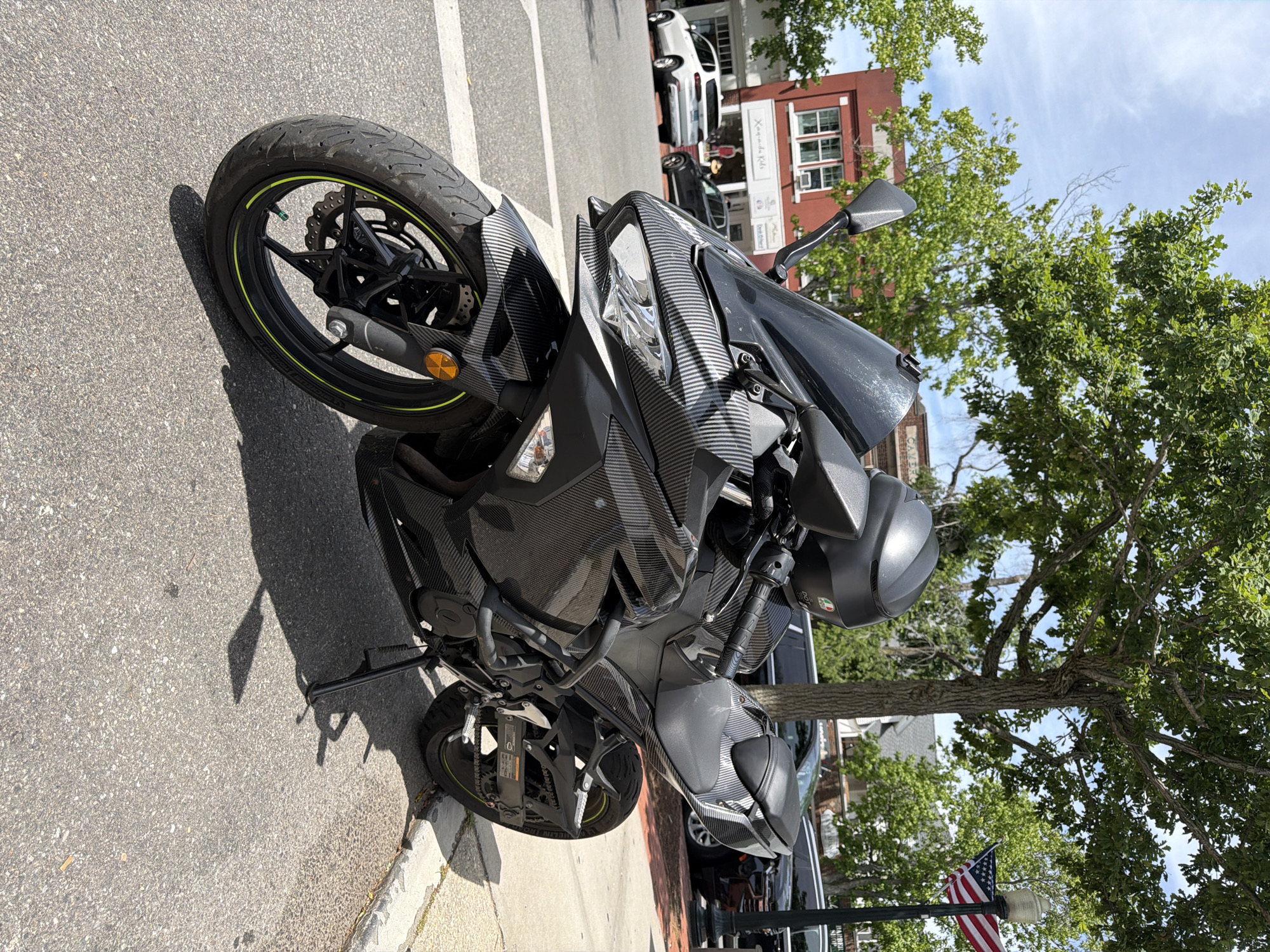 A black motorcycle, adorned with a carbon fiber wrap, is parked on a street in the Village of Southampton, USA. The motorcycle is the primary subject, positioned vertically in the frame, facing to the left. Its front wheel and part of the fairing are clearly visible, showing intricate carbon fiber detailing. The motorcycle is parked on an asphalt street with a sidewalk to its right.
In the background, a tree with lush green foliage partially obscures buildings. One building, appearing to be made of brick, has multiple windows. Across the street, other vehicles and buildings are visible, suggesting an urban or town setting. An American flag flies from a lamppost in the lower right portion of the image, near the sidewalk and a tree.
The scene appears to be set during the daytime, with bright sunlight casting shadows on the street, indicating it is likely a sunny day. The sky is visible at the top, with some clouds present. There is no clear indication of any specific activity or event taking place, other than the motorcycle being parked. The overall mood is calm and ordinary, reflecting a typical day in a village environment.
No discernible text is visible on the motorcycle or in the immediate surroundings, other than a small sticker on the motorcycle's mirror which is not legible.