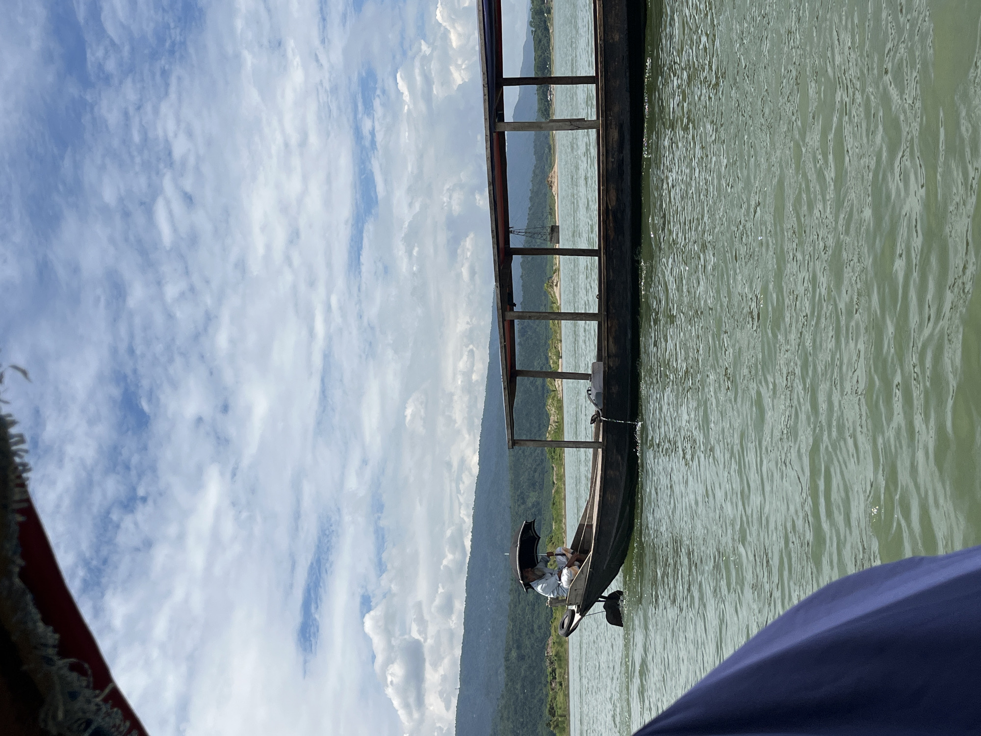 The image shows a wooden boat with a covered seating area on a calm river. A single person, wearing a light-colored shirt and white pants, sits in the front of the boat holding a black umbrella. The boat is moving, indicated by the wake behind it. In the background, there are lush green hills and a cloudy blue sky. The water is a greenish-blue color. The foreground shows a portion of another boat, covered in a dark blue fabric, suggesting the photo was taken from another vessel. The scene appears to be in a natural, outdoor environment. Without further context, pinpointing the exact location in Majai, India, is not possible solely from the visual information provided.