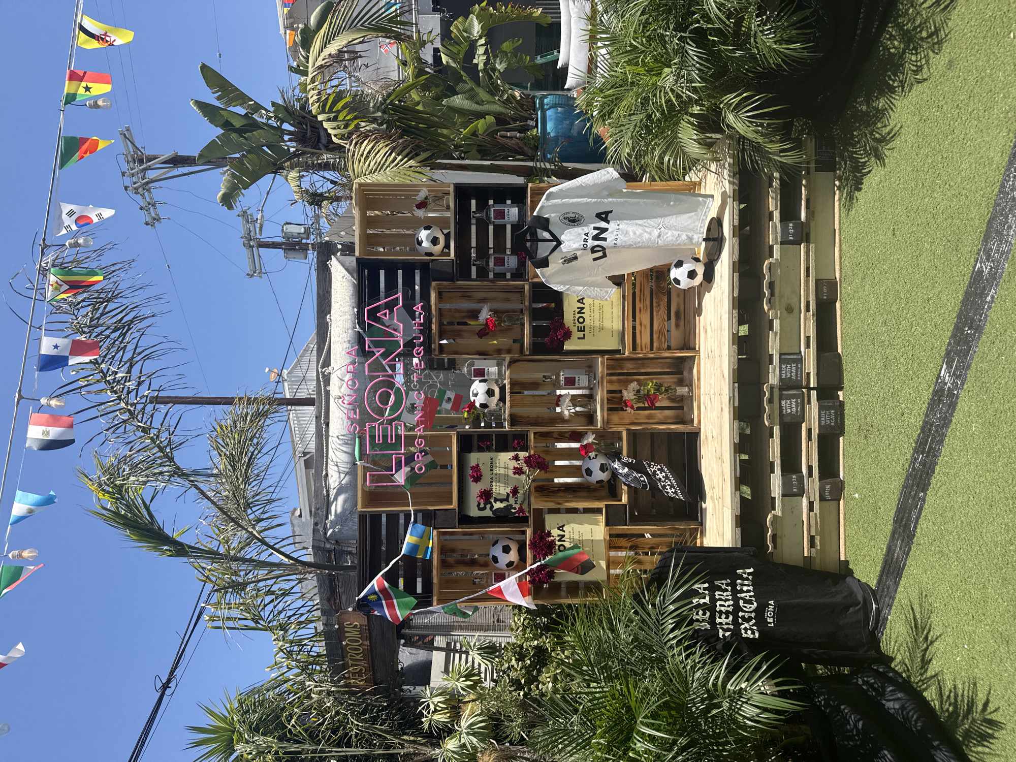 This is a medium shot, outdoor, eye-level, full-color photograph of a display featuring bottles of "Señora Leona Organic Tequila" and merchandise at a street-side venue in Santa Monica, California, United States of America. The display is set up against a weathered fence and includes various wooden crates, some stacked in a pyramid formation. Bottles of tequila, two soccer balls, and floral arrangements are placed within the crates. A white short-sleeved shirt with the "Señora Leona" logo and "Tequila" is hanging from a rack in the center of the display. A black t-shirt with white lettering reading "De La Tierra Exicana" and "Leona" is hung to the left of the display. Several national flags are strung across the top of the display. The ground in front of the display is covered with artificial turf, and lush green foliage and palm trees are visible around the scene. The sky is clear and blue. A sign for "RESTROOMS" is visible on the fence to the left of the display.