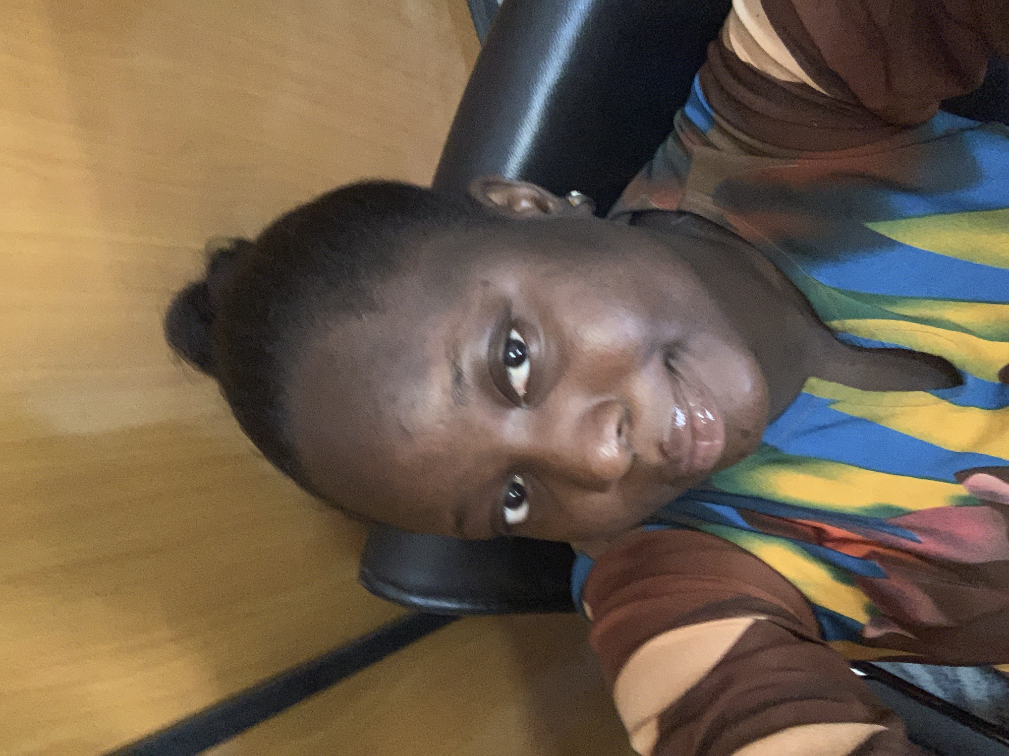 The image is a medium close-up selfie of a Black woman with dark brown skin and her hair pulled up in a bun. She is wearing a colorful shirt with abstract patterns of blue, yellow, brown, and red. She has a small hoop earring in her right ear. The background appears to be an indoor setting with a wooden wall and a black chair. The lighting is soft and appears to be from a natural source. The woman is looking directly at the camera with a slight smile. The overall tone of the image is casual and personal.