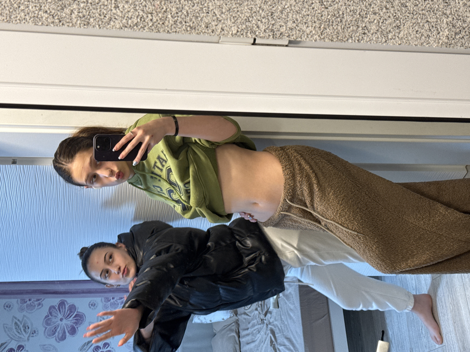 The image depicts a woman taking a selfie in a mirror. She is wearing an olive green sweatshirt that exposes her abdomen and tan sweatpants. Her hair is pulled back. The reflection shows a second woman wearing a black puffer jacket and white pants, standing in what appears to be a bedroom with floral wallpaper. The location is unknown, and there's no information to determine if this scene is taking place in Sofia, Bulgaria.