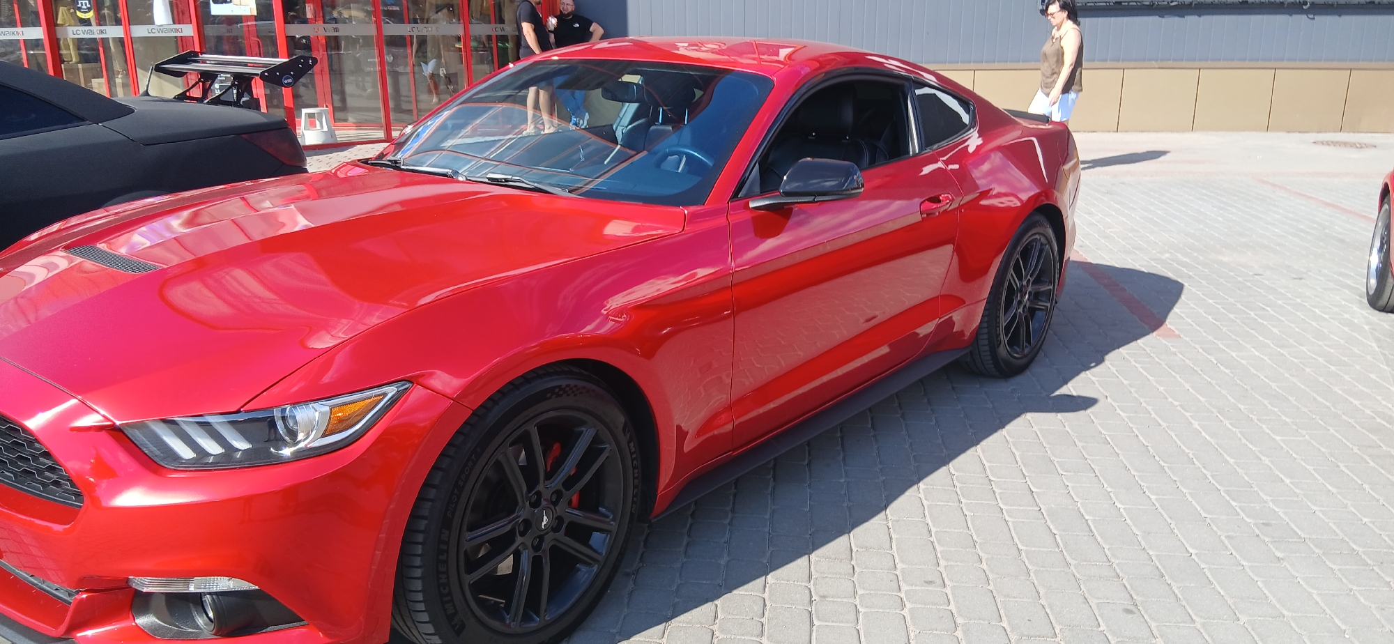 A bright red Ford Mustang is parked on a paved lot, with a black car with a spoiler behind it. The Mustang is a coupe with black rims and red brake calipers. The scene appears to be outdoors during the daytime, with the sun casting shadows on the ground. The background shows a building with large windows and signage. There are two people visible: a man standing near the building and a woman walking in the background. The building appears to be a retail establishment, with text that is partially visible reading "LC WAIKIKI". The overall mood seems casual, possibly a car show or a gathering of car enthusiasts.