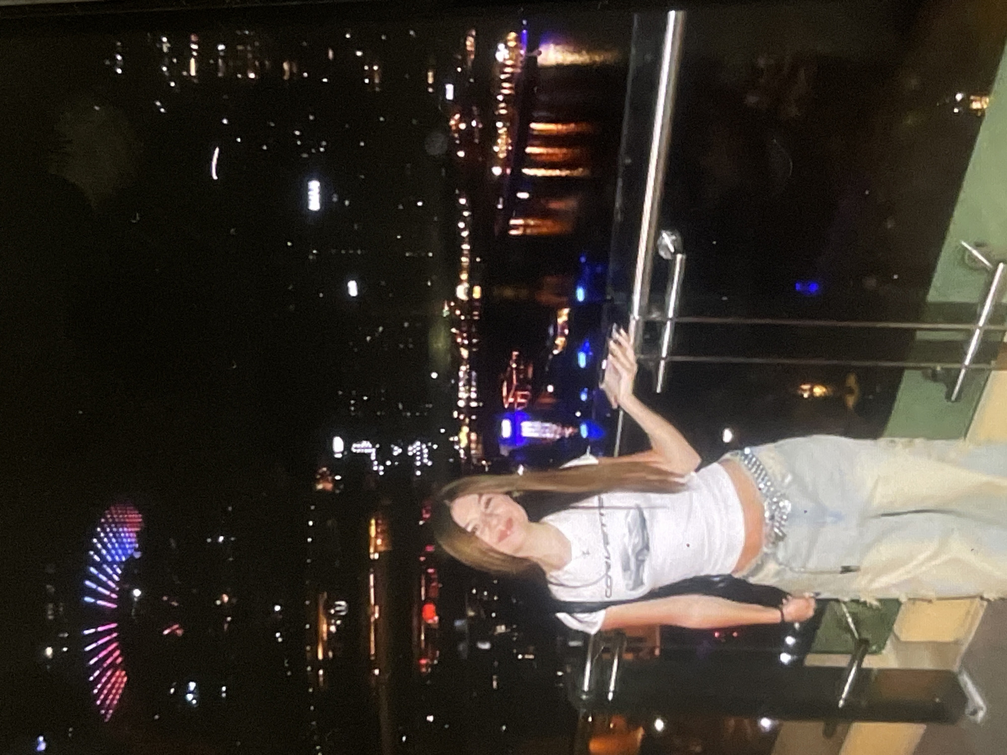 An image, initially rotated, depicts a young woman posing on a balcony or elevated platform at night, overlooking a bustling city skyline in Poltava, Ukraine. The scene is illuminated by countless artificial lights from buildings and reflections on a body of water, likely a river, stretching into the distance.
The subject is a woman with long, straight brown hair, wearing a white short-sleeved top and light-colored, possibly ripped, jeans. A prominent, ornate silver belt with studs or chains cinches her waist. She has a faint smile and appears relaxed, with one arm raised and bent. Her white top features a dark graphic and visible text that reads "CORVETTE," accompanied by an outline of a car.
In the background, the dark night sky contrasts with the bright, warm-toned lights of various buildings and streetlights. To the left, a large Ferris wheel glows with distinctive red and blue lights, serving as a prominent landmark. The foreground shows parts of a modern railing, suggesting a high-rise vantage point. The overall impression is one of an urban nightscape, captured in a casual portrait. The image quality is somewhat grainy, consistent with low-light photography.