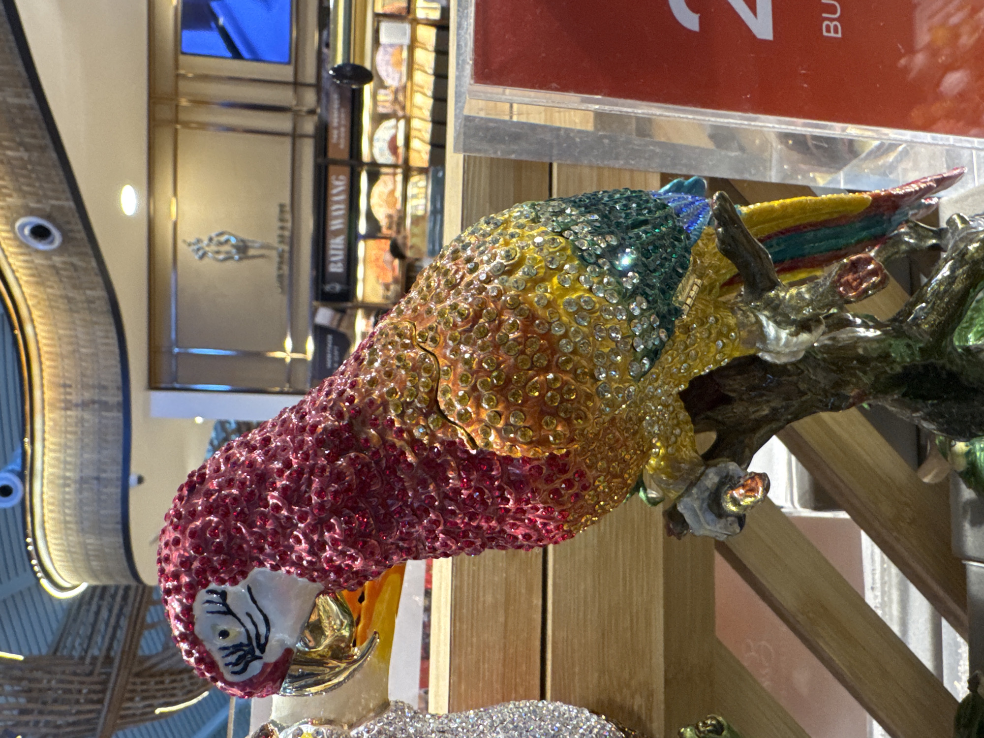 A close-up, canted, medium-shot shows a bejeweled parrot figurine perched on a branch. The parrot's head is predominantly red, transitioning to orange, yellow, and finally green near the top of its head. Its tail feathers are blue, yellow, and red. The branch the parrot is perched on is painted in shades of green and gold.
In the blurred background, one can discern parts of a building interior, possibly a shopping mall or a store. A sign "Batik Wayang" is visible. Based on this sign, it is likely the location is somewhere in the city of Kuta, Indonesia, given Batik Wayang is a store chain that specializes in Indonesian batik art and souvenirs.