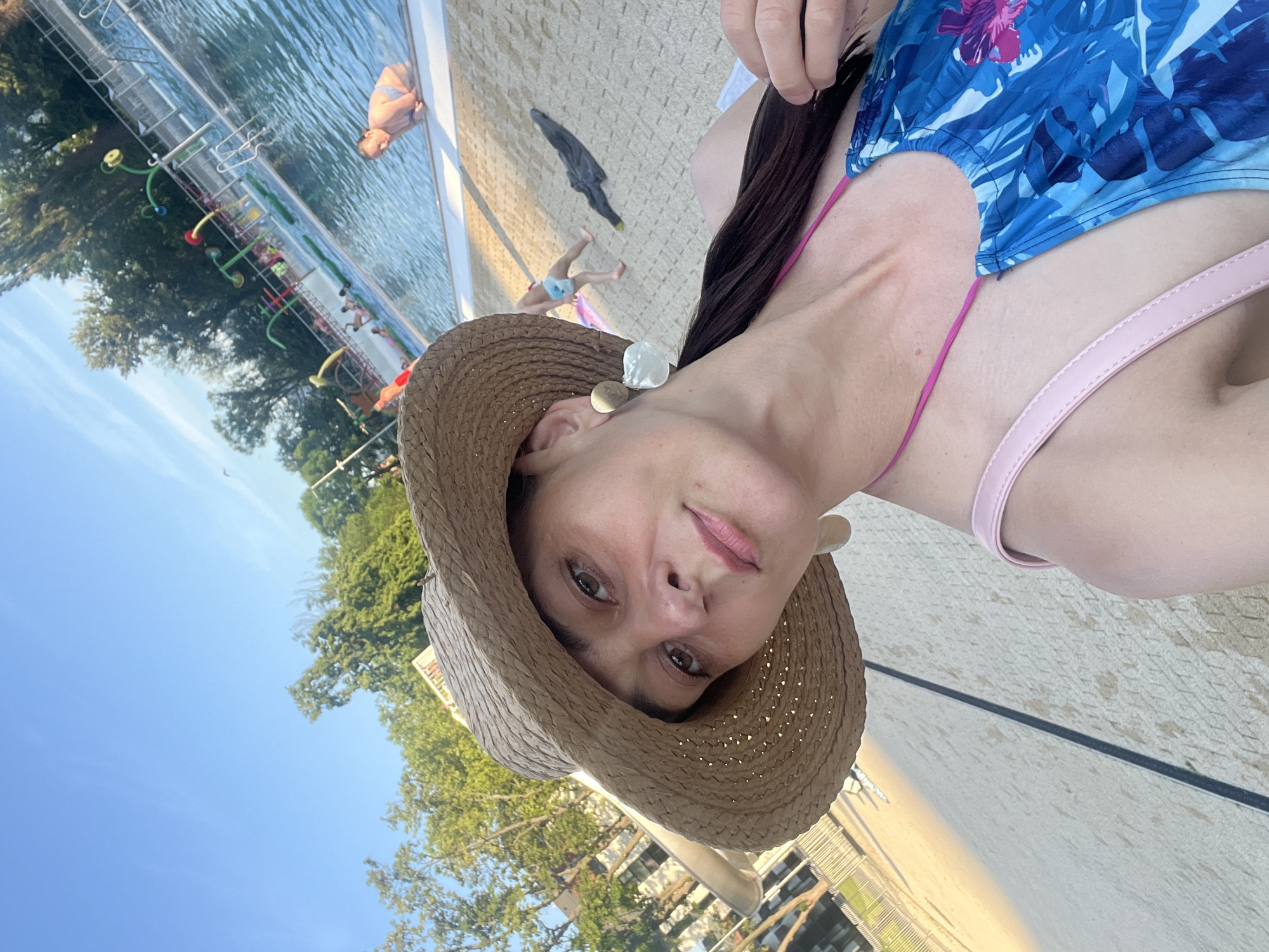 A woman in a blue floral swimsuit and a straw hat takes a selfie at a public pool. She is wearing large, white earrings and has a pink purse strap visible. The pool is filled with people, some swimming and others relaxing on the edge. The background shows a sunny day with trees and a clear blue sky.