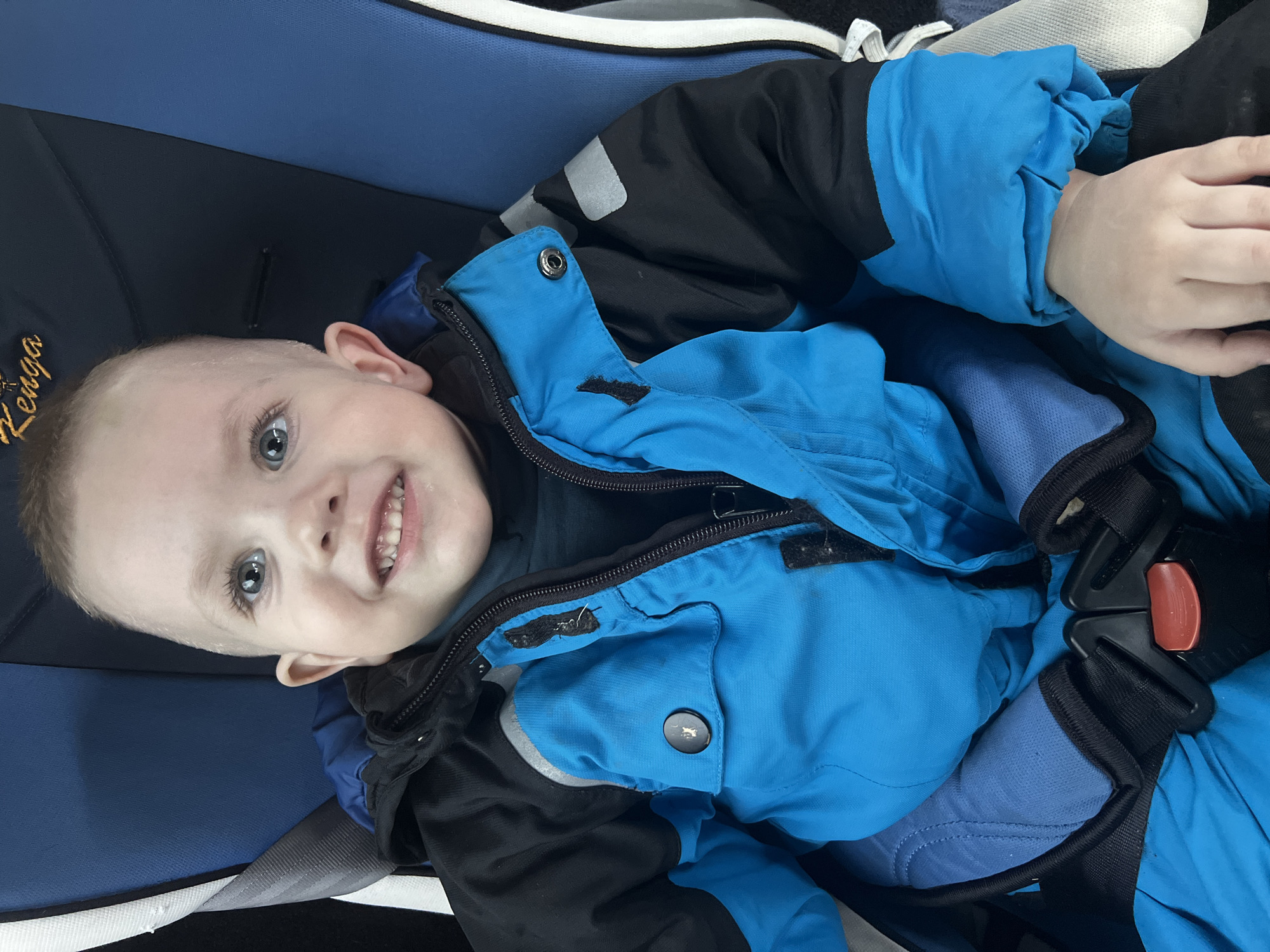 A young child is seated in a blue and black car seat within the interior of a vehicle. The child, with light-colored eyes and short light hair, is smiling directly at the camera, displaying several teeth and exhibiting some moisture around the mouth area. The child is wearing a two-tone blue and black winter jacket with reflective patches on the shoulders, over a dark-colored inner garment. The car seat features a headrest with the word "Zenga" embroidered in gold lettering and a five-point harness system securing the child, with a visible red release button on the buckle. A grey and white striped fabric is visible to the child's right, and a grey seatbelt strap is visible to the child's left. The scene is located in Chaikovsky, Russia.