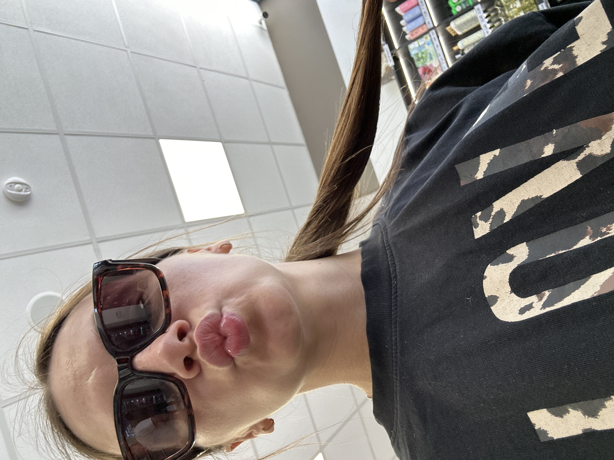 A young woman with brown hair pulled back in a ponytail, wearing sunglasses and a black t-shirt with a cow print design, makes a kissing face towards the camera. She is indoors, possibly in a store, with a tiled ceiling and fluorescent lighting visible above. Shelves stocked with various products are blurred in the background. The scene is captured from a low angle, with the woman's face and upper body dominating the foreground. The image does not provide any specific information about the location within the city of Donji Pobori, Montenegro.