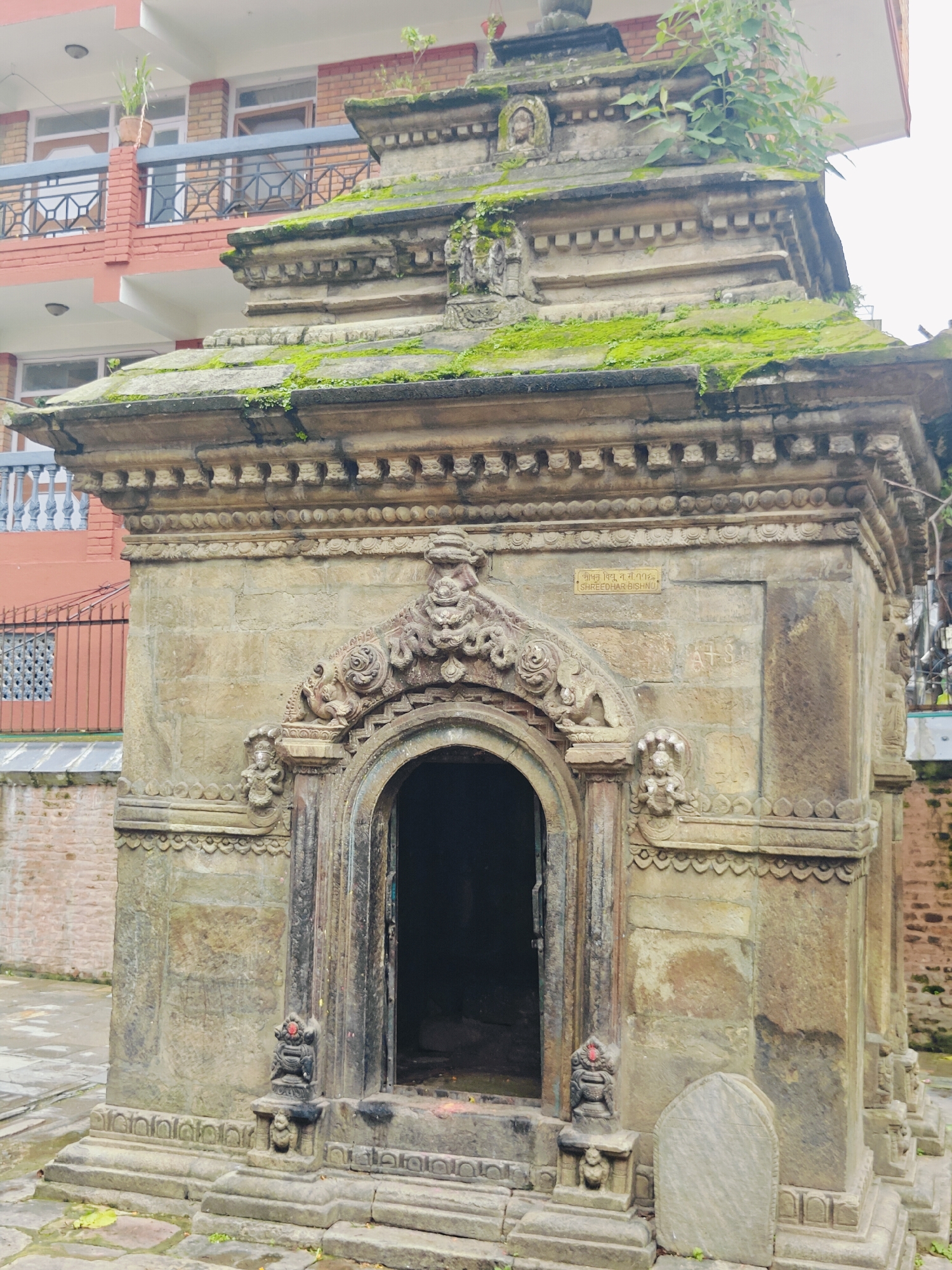 The image shows a weathered stone temple structure, likely an ancient shrine, located in Kirtipur, Nepal. The temple is ornate, with intricate carvings around its arched doorway and along its roofline. Moss and some vegetation have grown over parts of the structure, indicating its age and exposure to the elements. A metal railing and a portion of a brick building are visible in the background, suggesting the temple is situated within a populated area. The ground is paved with stone and appears to be slightly damp, hinting at recent rain. No people are present in the scene.