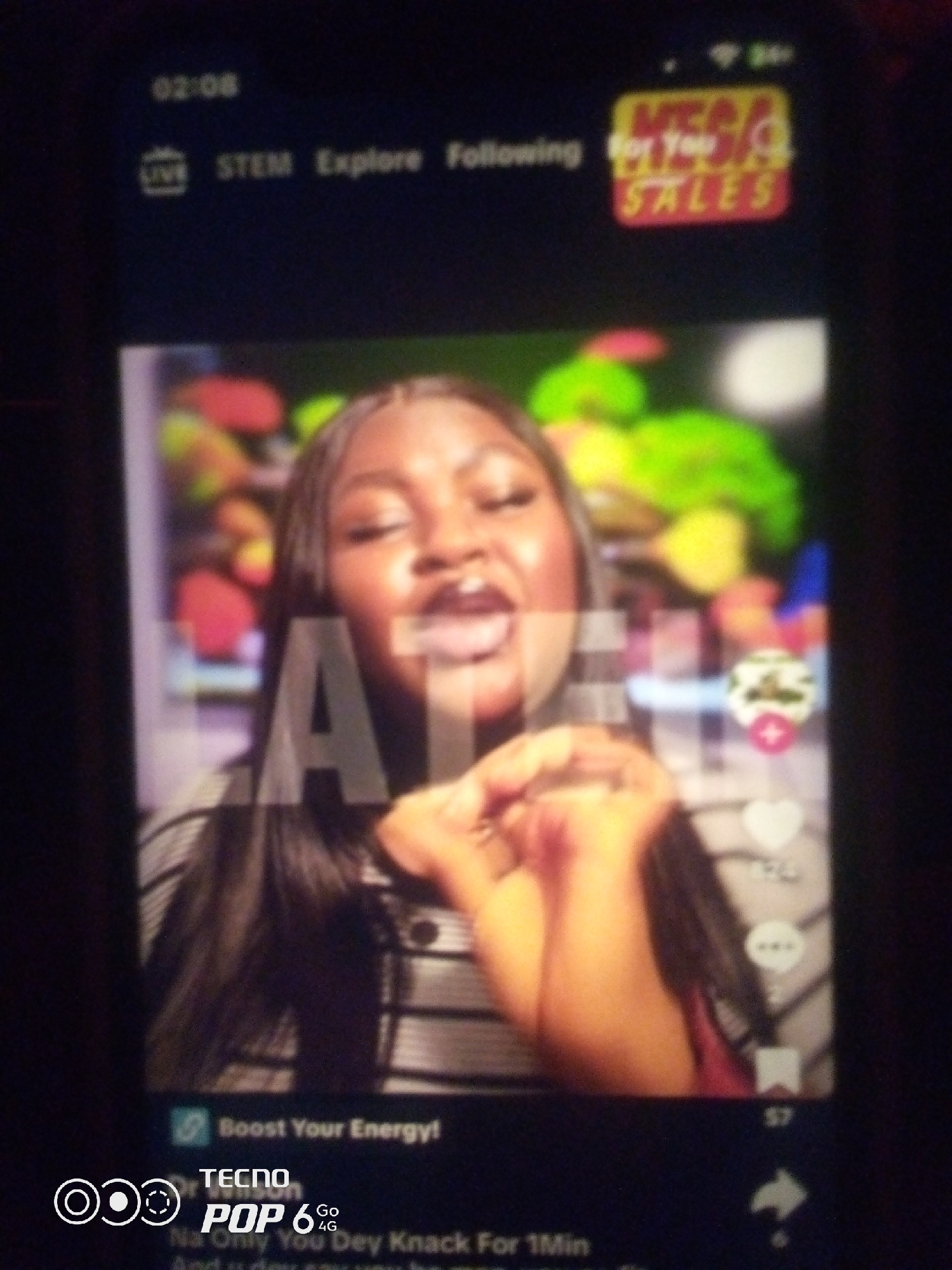 The image shows a close-up, blurry view of a person's face on a mobile phone screen. The person appears to be a young Black woman with her eyes closed and her mouth open. She is wearing a striped shirt and has long, dark hair. In the background, there are colorful, out-of-focus shapes that might be fruit or vegetables. Overlaid on the image are various graphic elements typical of a social media app, including icons for "LIVE," "STEM," "Explore," and "Following." There is also a "For You Sales" banner in the top right corner and text at the bottom that reads "Boost Your Energy!" and "TECNO POP 6 Go 4G." The specific location of Howayi, Nigeria, cannot be determined from the visual information provided in the image.