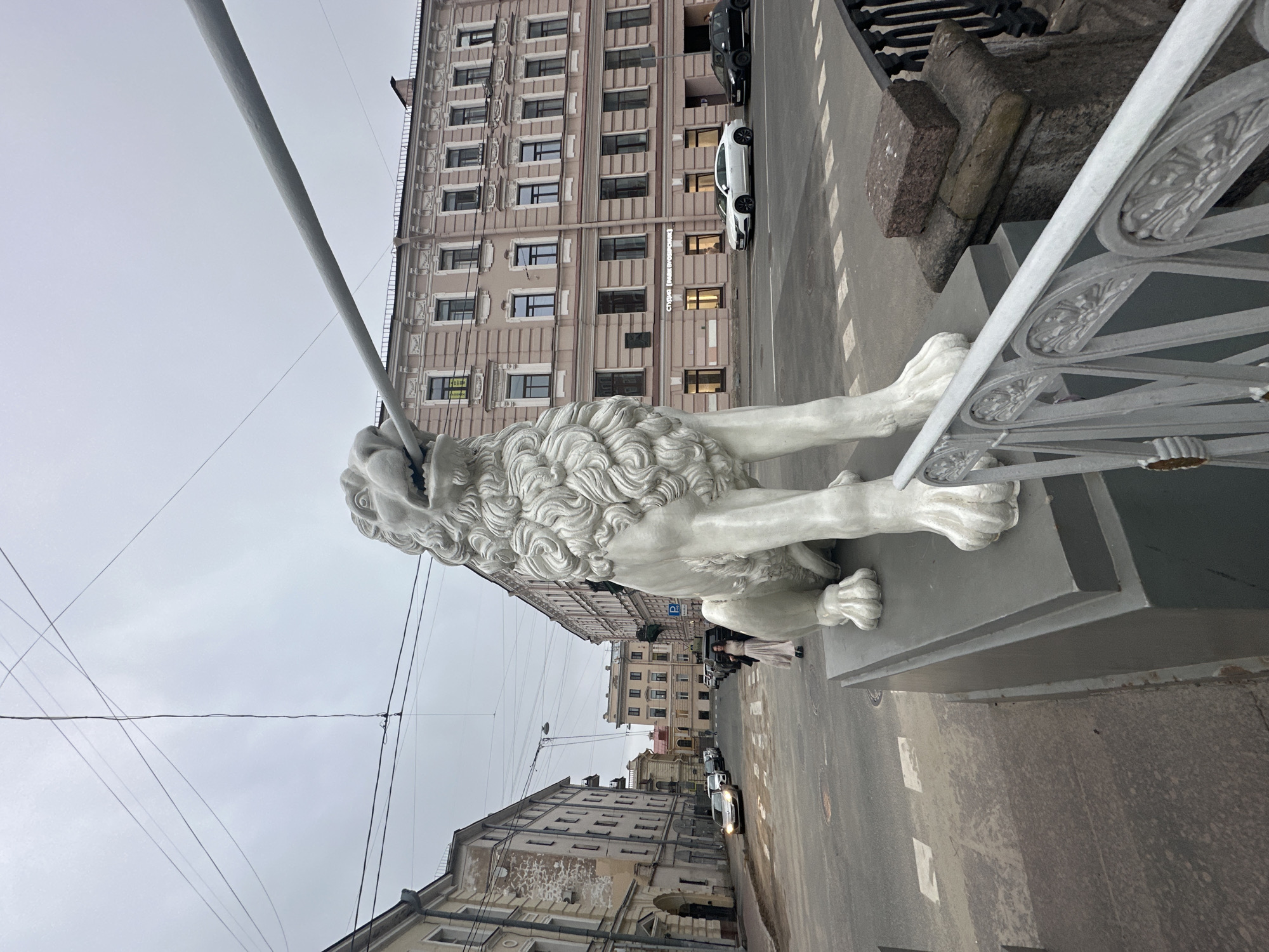 The image shows a statue of a lion on a bridge in Saint Petersburg, Russia. The statue is white and detailed, positioned with its front paws extended onto the bridge's base. The bridge features decorative metal railings. In the background, there is a multi-story building with numerous windows and a street with cars and pedestrians. The sky is overcast.