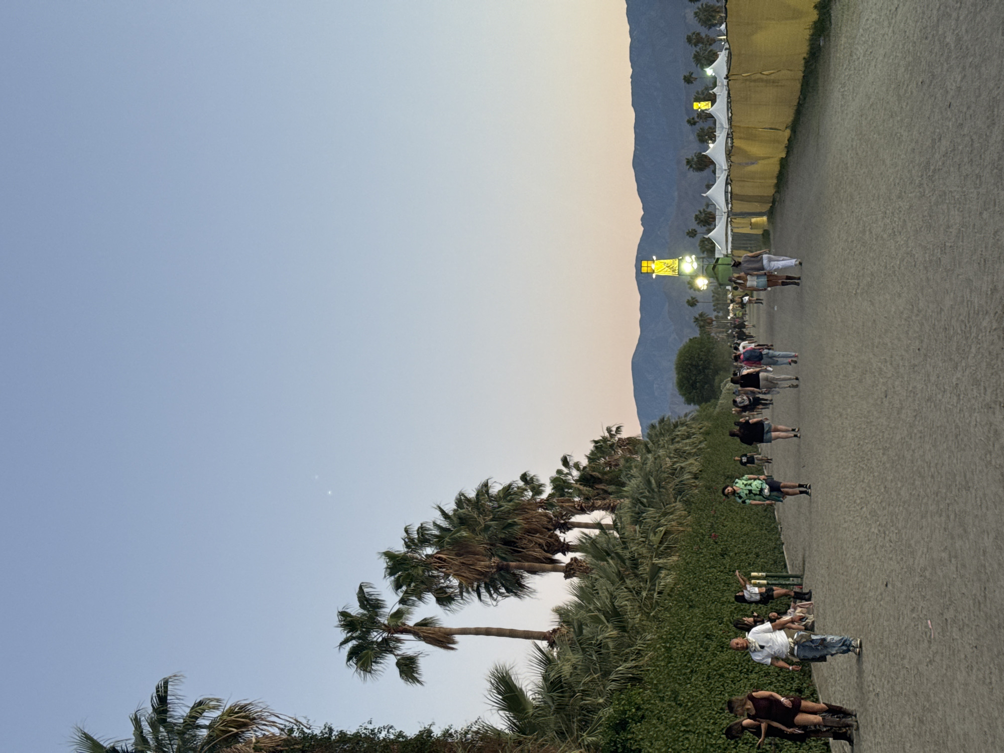 The image depicts a scene in Indio, California, United States, during what appears to be a festival or large event. A large group of people are walking on a sandy or dirt path alongside palm trees and green vegetation. The background features tents, fencing, mountains, and a clear sky with a gradient of blue and orange light suggesting either sunrise or sunset. The people are dressed in casual clothing, and their actions suggest they are moving between locations within the event grounds.