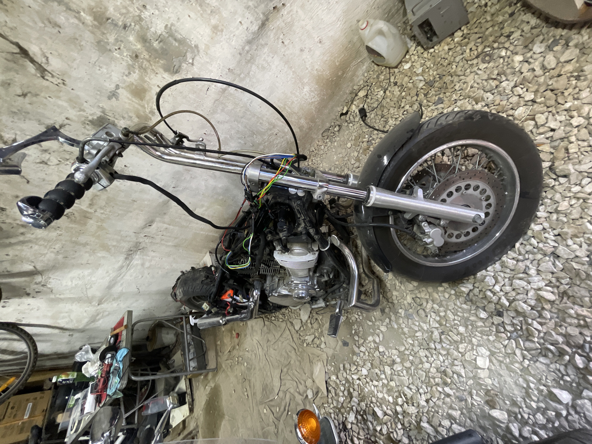 An indoor scene shows a partially disassembled motorcycle, likely a cruiser or chopper, lying on its left side on a garage floor. The motorcycle features a large, chromed V-twin engine with visible cooling fins and exhaust pipes. Its front end includes a long, chromed fork assembly, a front wheel with a disc brake and spoked rim, and handlebars equipped with grips and brake/clutch levers. Various colored wires (red, green, yellow, blue, black) are exposed around the steering head and engine, suggesting ongoing repair or customization work.
The floor beneath the motorcycle is a mix of rough concrete and light-colored gravel, with a beige tarp or fabric laid out under the engine area. In the background, against a rough concrete wall, several items are visible: a white plastic jug, a gray electronic box, and a metal shelving unit cluttered with tools, rags, and other miscellaneous parts. Part of a bicycle wheel is visible on the far left, and an orange turn signal lens is seen in the bottom right corner, possibly belonging to another part of the motorcycle or another vehicle. The scene conveys an active workshop environment, with the motorcycle in a state of maintenance or reconstruction. No people are visible.
