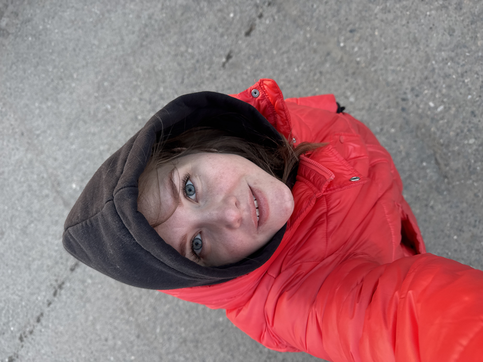 An overhead photograph captures a young person looking directly up at the camera. The subject has fair skin, striking blue eyes, and light brown hair partially visible beneath a dark grey or black hoodie. They are wearing a vibrant red puffer jacket, which contrasts sharply with the muted grey background. A gentle, friendly smile is present on their face, and their eyes convey engagement.
The background consists of a textured, light grey surface, appearing to be asphalt or concrete, which suggests an outdoor setting, possibly a street or sidewalk in Горный, Russia. The lighting is diffused and even, indicating either an overcast day or a location shielded from direct sunlight. The perspective of the shot, taken from above, emphasizes the subject looking upwards. No visible text is present in the image.